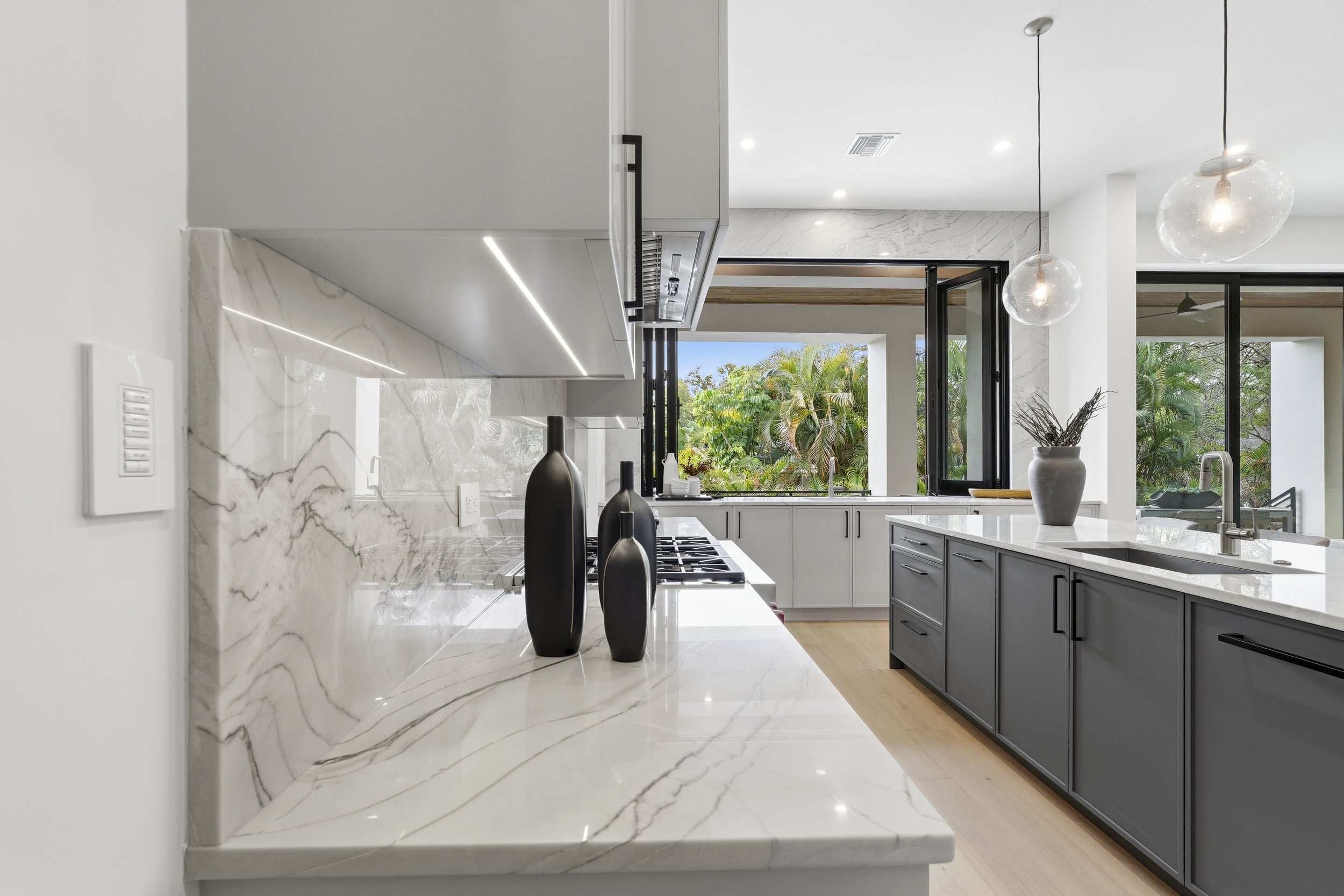 Modern kitchen with white marble countertops and backsplash, black pendant lights, and large window showing greenery outside.