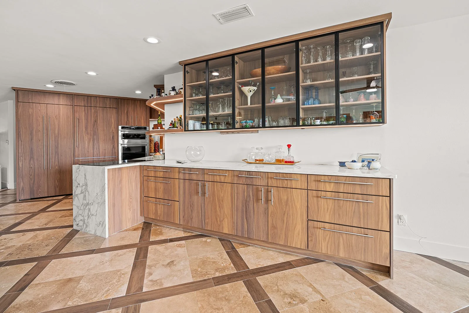 Modern kitchen with wooden cabinets, marble countertops, and glass-front upper cabinets filled with glassware and decorative items.