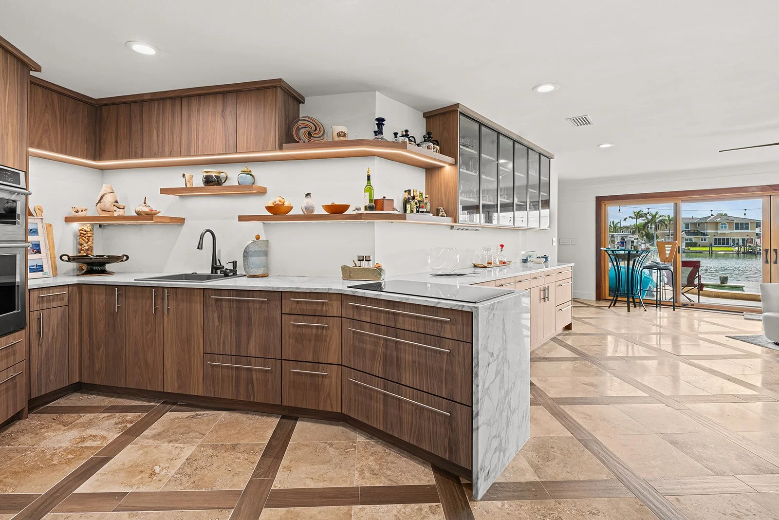 Modern kitchen with wooden cabinets, marble countertop island, open shelves with decorative items, and a view of a waterfront patio and houses outside.