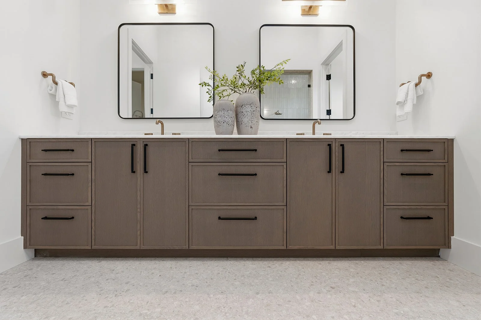 A modern bathroom vanity with a double sink, two mirrors, and two decorative vases with green foliage on top.