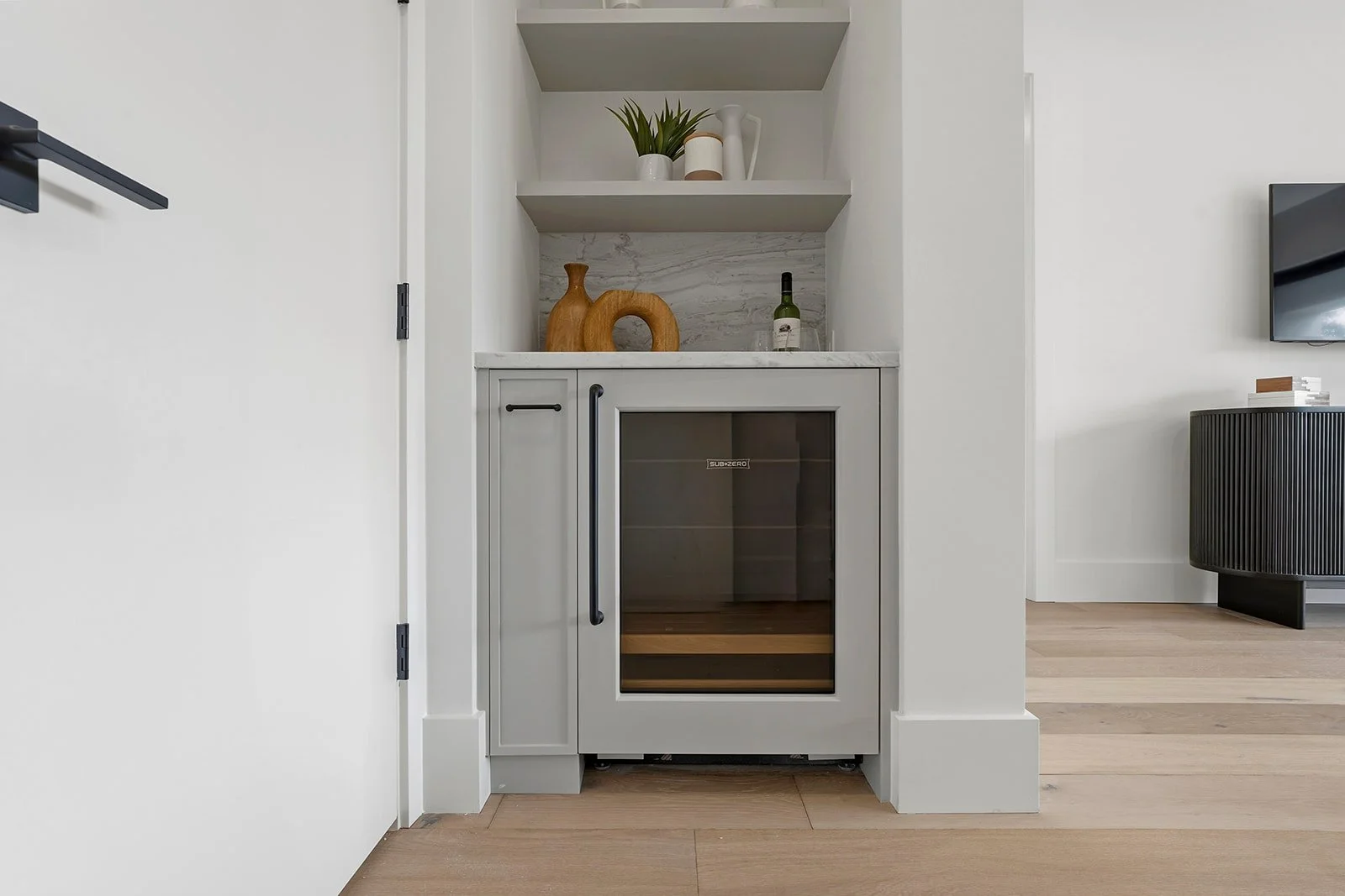 A small portable wine cooler with a gray frame and glass door sitting in a corner with decorative wooden and ceramic vases and a green bottle on top. There are white walls and a doorway, with part of a black speaker or cabinet and a black TV monitor 