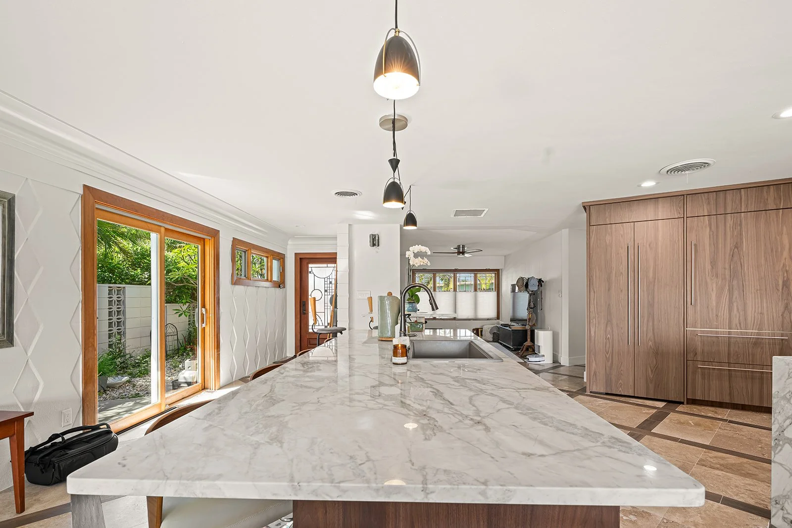 Interior view of a modern kitchen with a marble island, wooden cabinets, sliding glass door, and pendant lights.