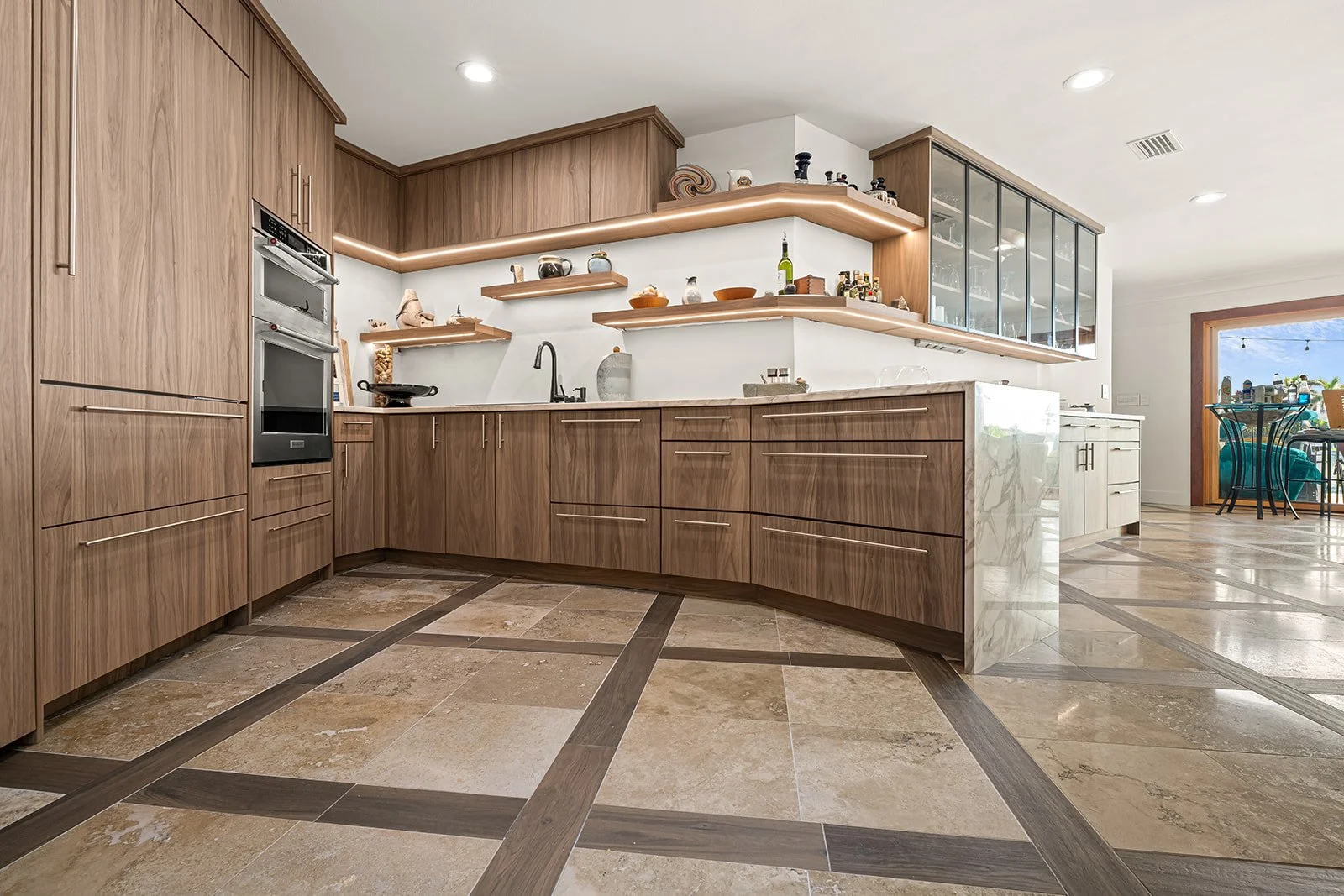 Modern kitchen with wood cabinets, beige tiled floor, and open shelves with decorative items. Brightly lit with natural light from a large window or door in the background.