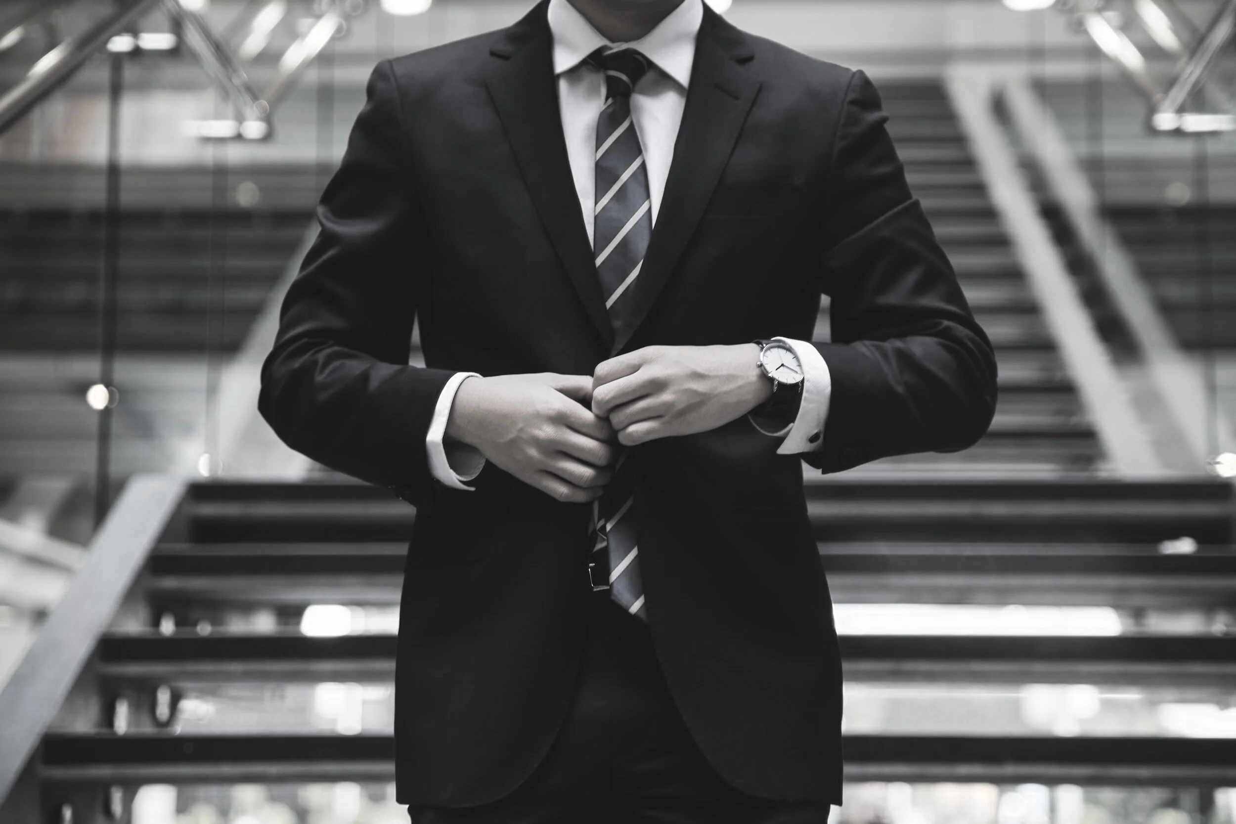 A person wearing a black suit, white shirt, and striped tie, standing on a staircase in an urban setting, with their arms crossed and a watch visible on their left wrist.