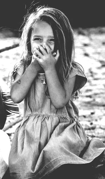 Gina as a young girl sitting outdoors, smiling and covering her mouth with her hand,