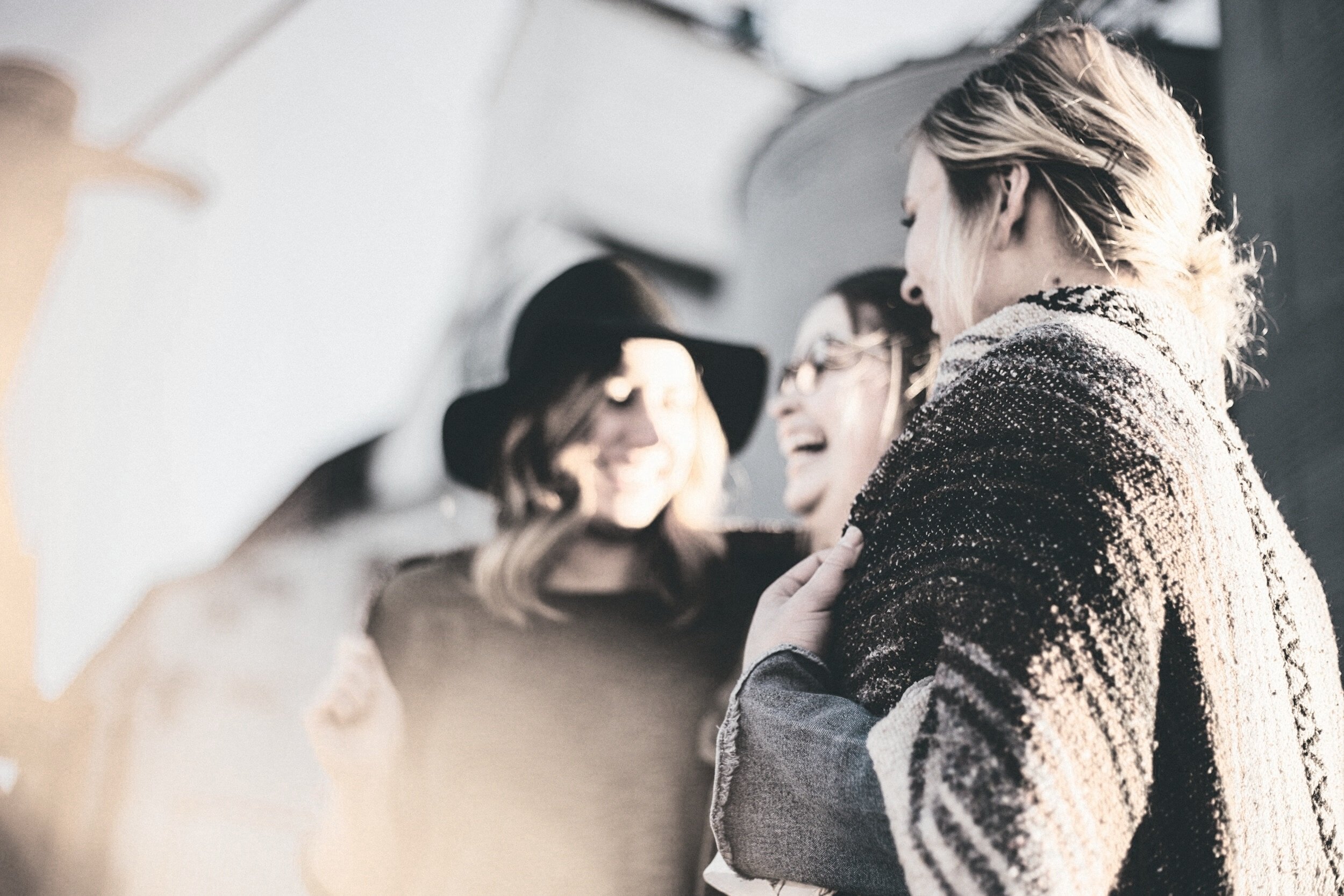 A group of women smiling and embracing each other, dressed warmly in jackets and scarves, in an outdoor setting.