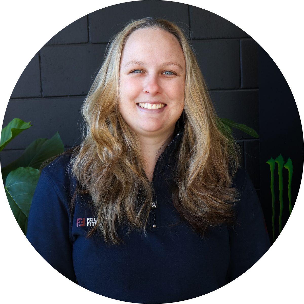 A smiling woman with long, wavy blonde hair wearing a black jacket with a small logo, standing in front of a black brick wall with green plants.