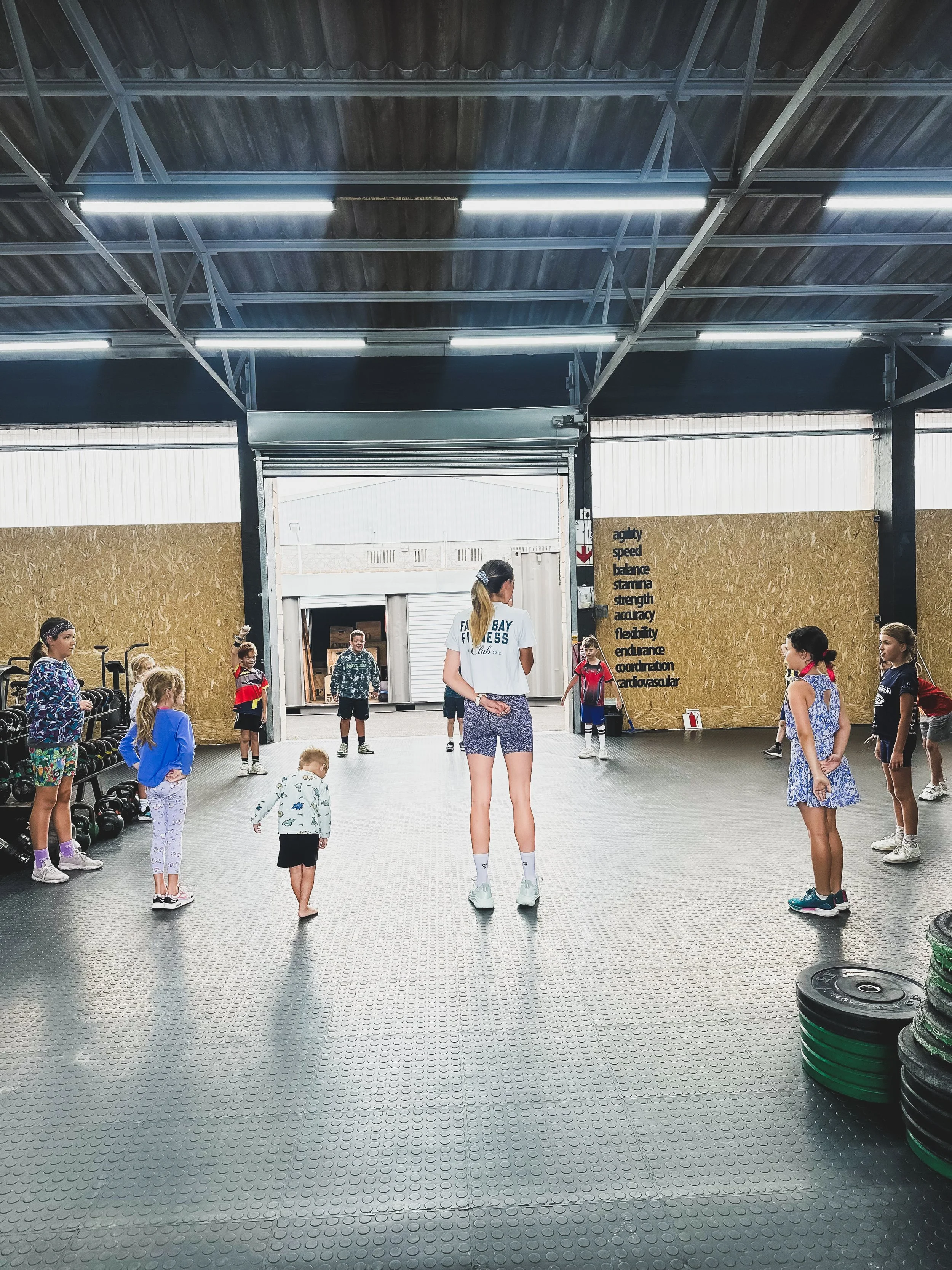 A fitness class taking place in an industrial-style gym with children and an instructor in the center, standing on a rubber flooring with weight plates stacked on the side and a wall with motivational words to the right.