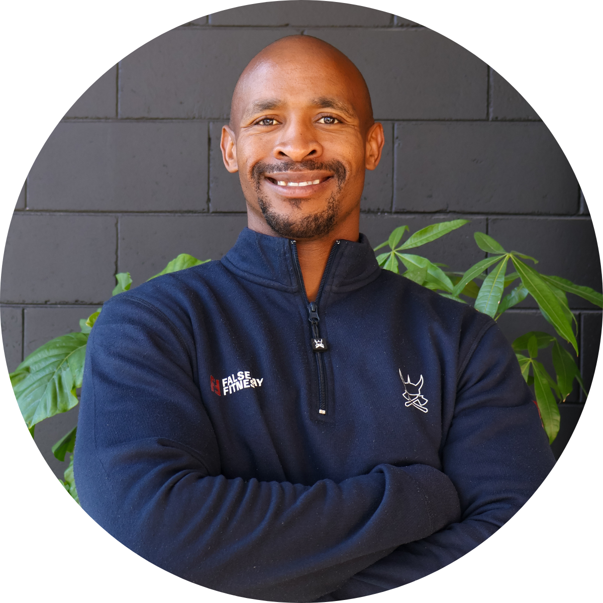 A smiling man with a goatee and bald head, wearing a navy Blue zip-up sweatshirt with the embroidered logo of a fitness company, standing in front of a gray brick wall with green plants.