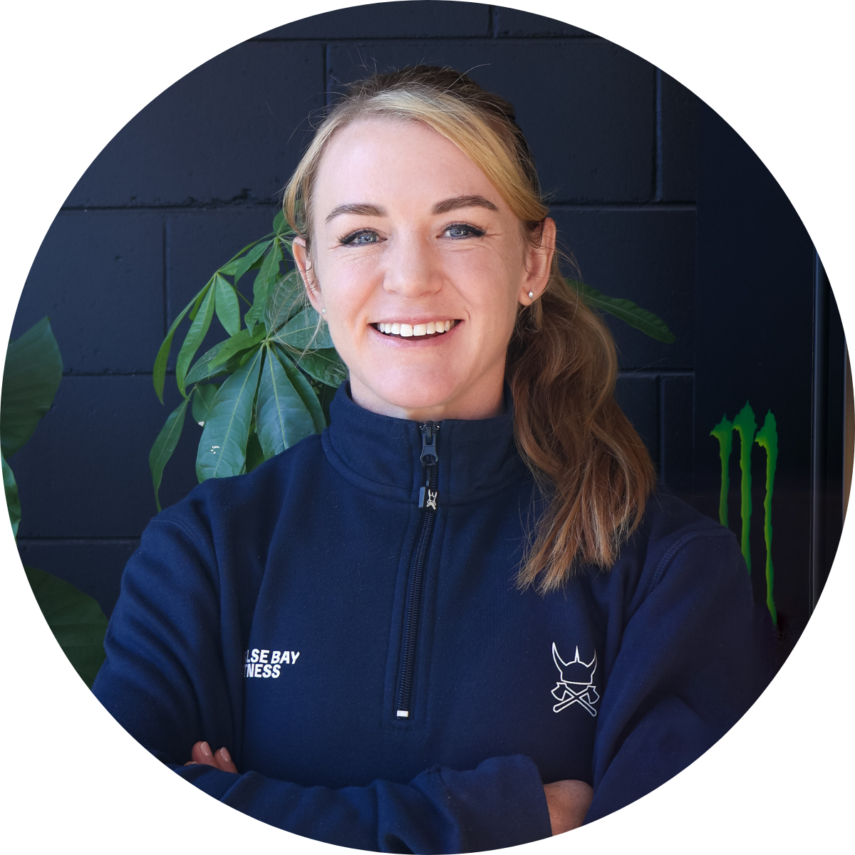 A woman with long, wavy light brown hair smiling confidently, wearing a navy blue zip-up jacket with a logo on the left side, standing in front of a black brick wall and green decorative plants, with a Monster energy drink logo in the background.