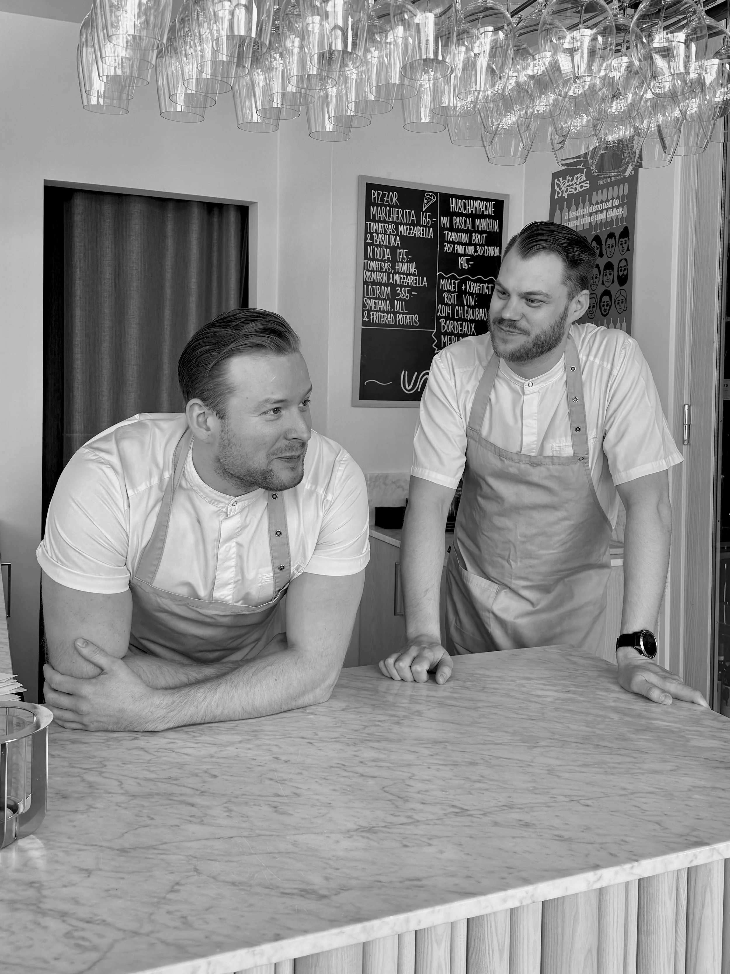 Two men in white shirts and aprons sitting at a marble countertop in a restaurant, engaging in conversation. There is a glass container on the left and a blackboard menu behind them.