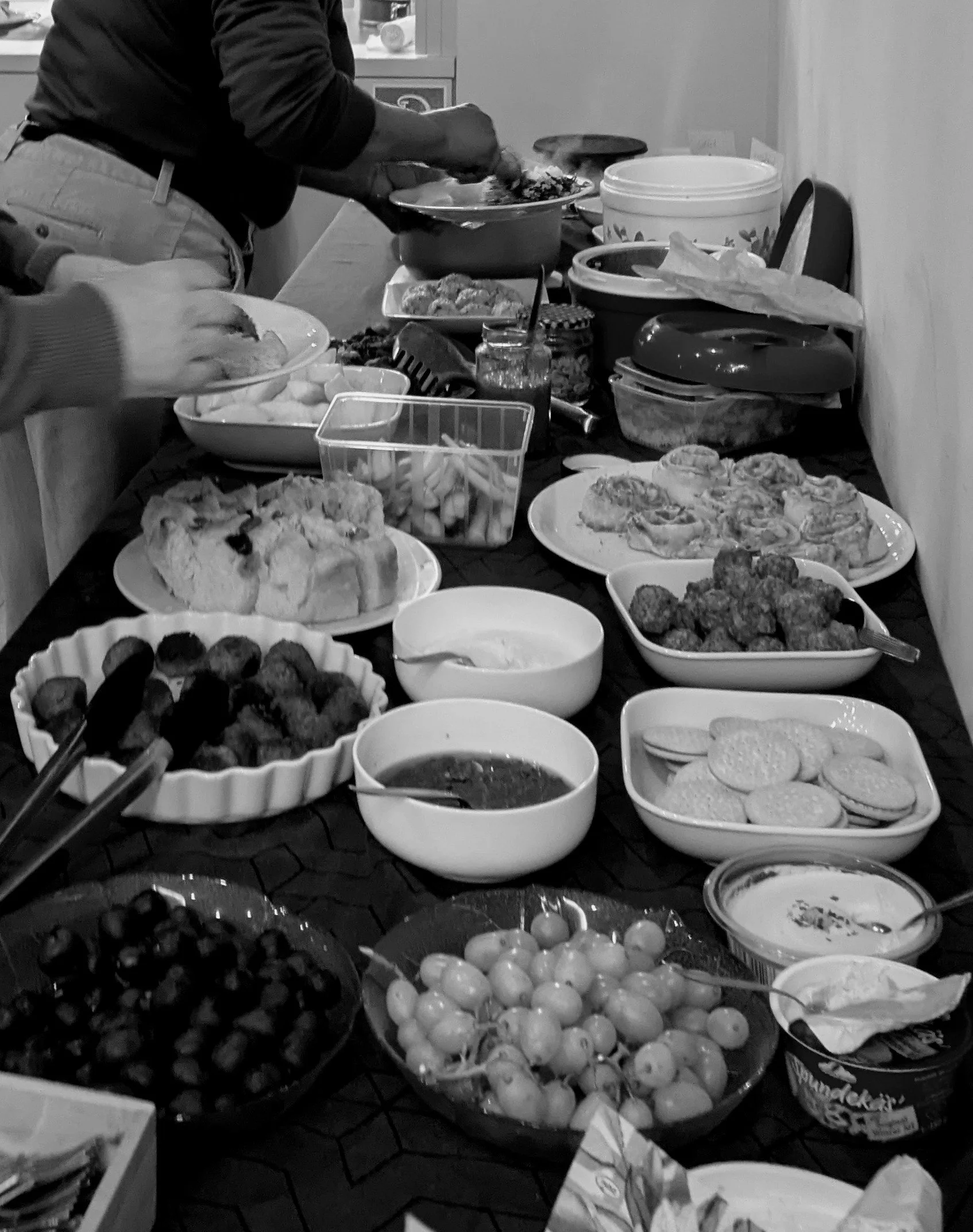 A table set with various dishes for a meal, including grapes, cookies, meatballs, bread, and salads, with people serving food.