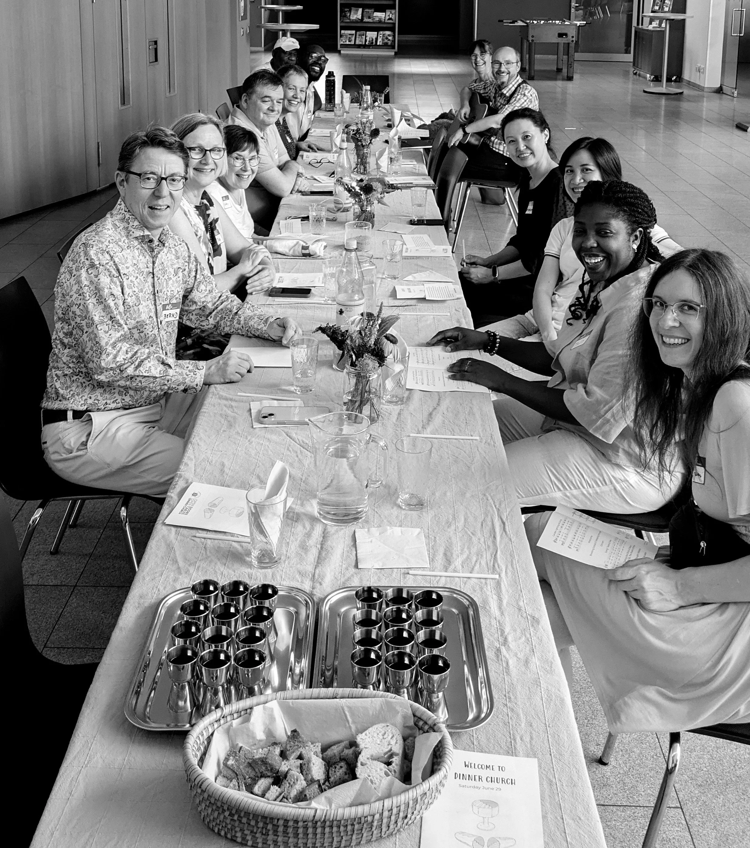 A black and white photo of a large group of people sitting around a long table, smiling for the camera. The table has glasses, plates, trays with shot glasses, and a basket of bread and pastries. There are also flowers and papers on the table.