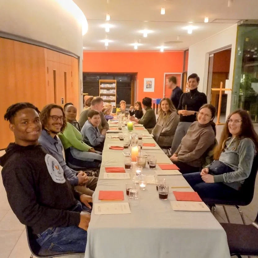 Group of people sitting around a long dinner table in a restaurant or banquet hall, smiling and enjoying a meal together, with candles and drinks on the table.