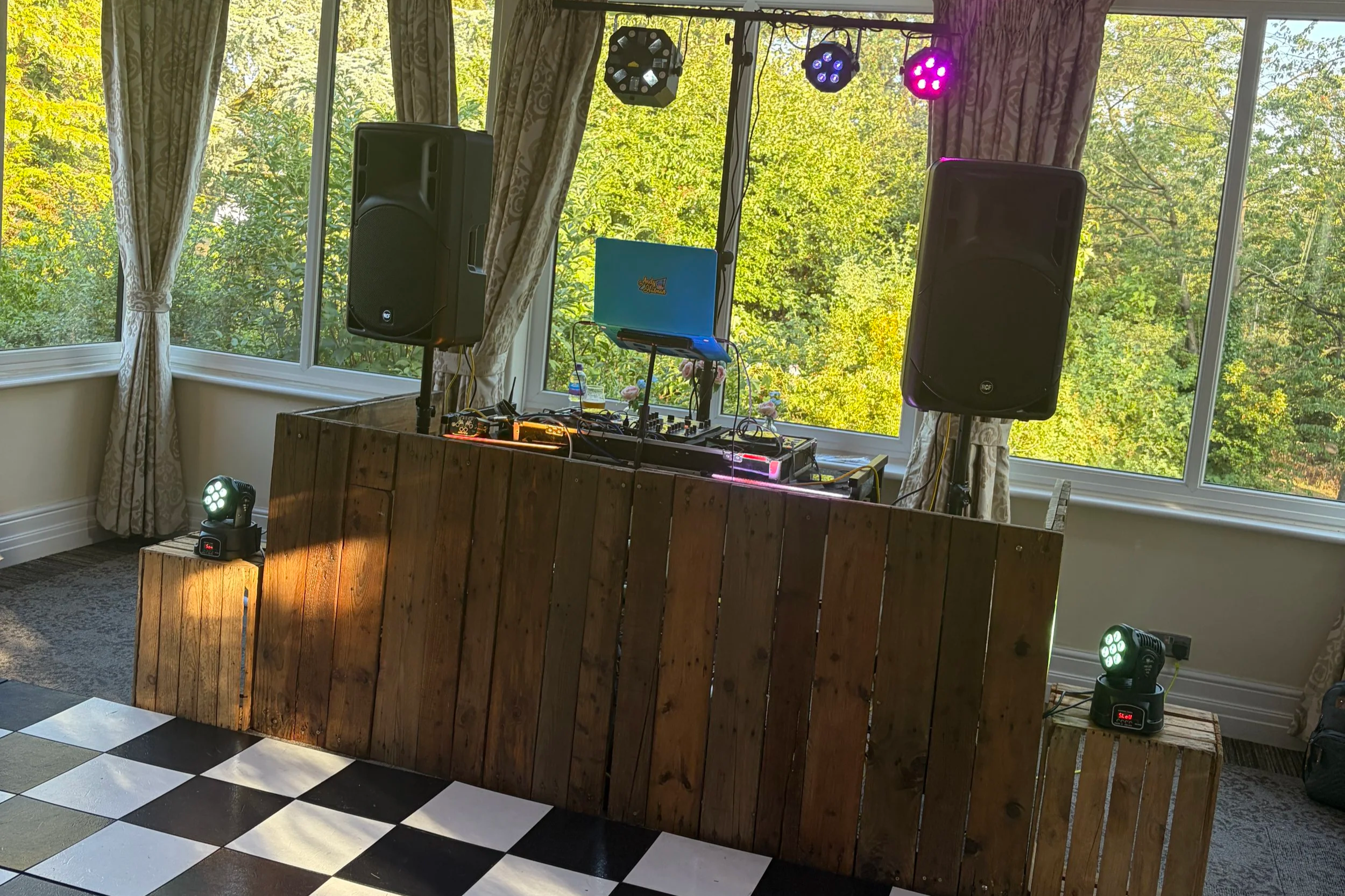 Indoor DJ setup with speakers and lighting beside large windows overlooking trees. Used for wedding hire