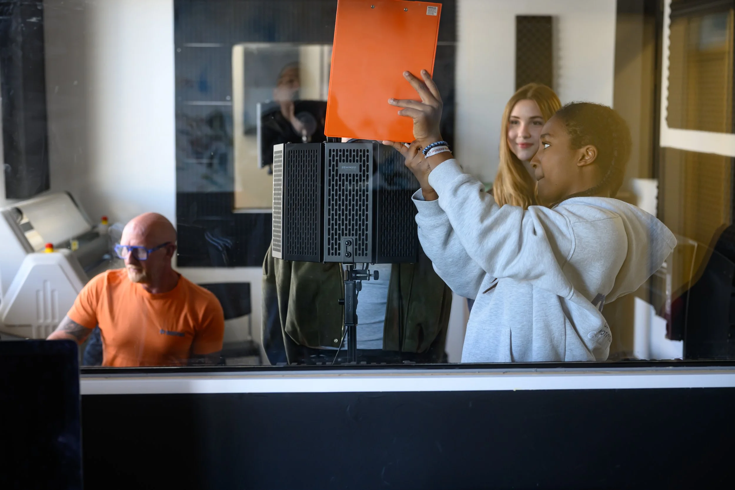 Drei Menschen in einem Raum, eine Person mit dunklem Haar und eine andere mit braunen Haaren beobachten eine Person, die ein orangefarbenes Gerät hochhält, durch eine Glasscheibe. Es gibt technische Geräte im Raum, die auf einen Studio- oder Forschun