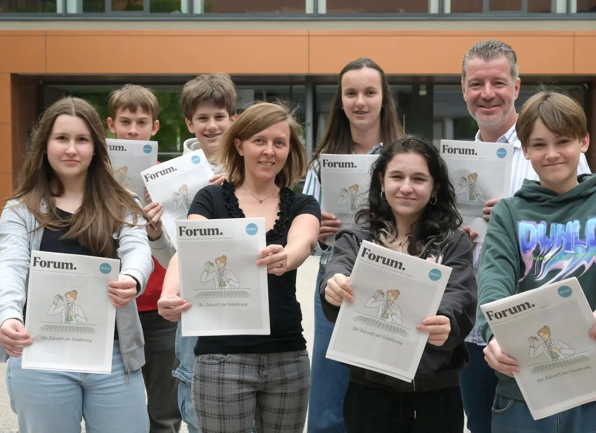 Eine Gruppe von Jugendlichen und einem Erwachsenen, die draußen vor einem modernen Gebäude stehen und Broschüren halten. Die Broschüren tragen den Titel »Forum« und haben eine Illustration einer Person, die etwas liest.