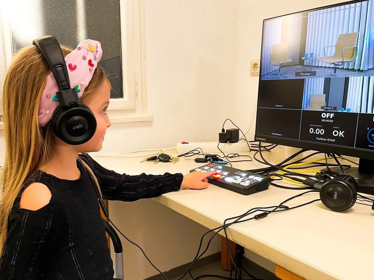 Ein junges Mädchen mit Kopfhörern sitzt vor einer Videoaufnahmetafel in einem Raum mit weißer Wand und kleiner Fenster, während sie eine Fernsehbildschirm überwacht.