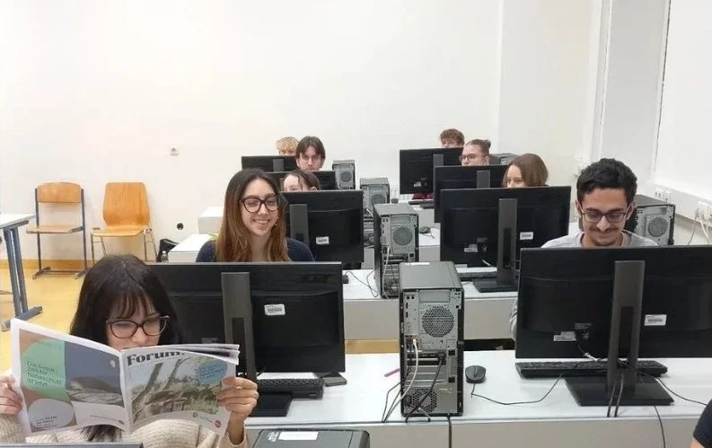 Mehrere Schüler in einem Computerraum, einige schauen auf Computerbildschirme, eine Person liest eine Zeitschrift, alle sitzen an Tischen mit Computern.
