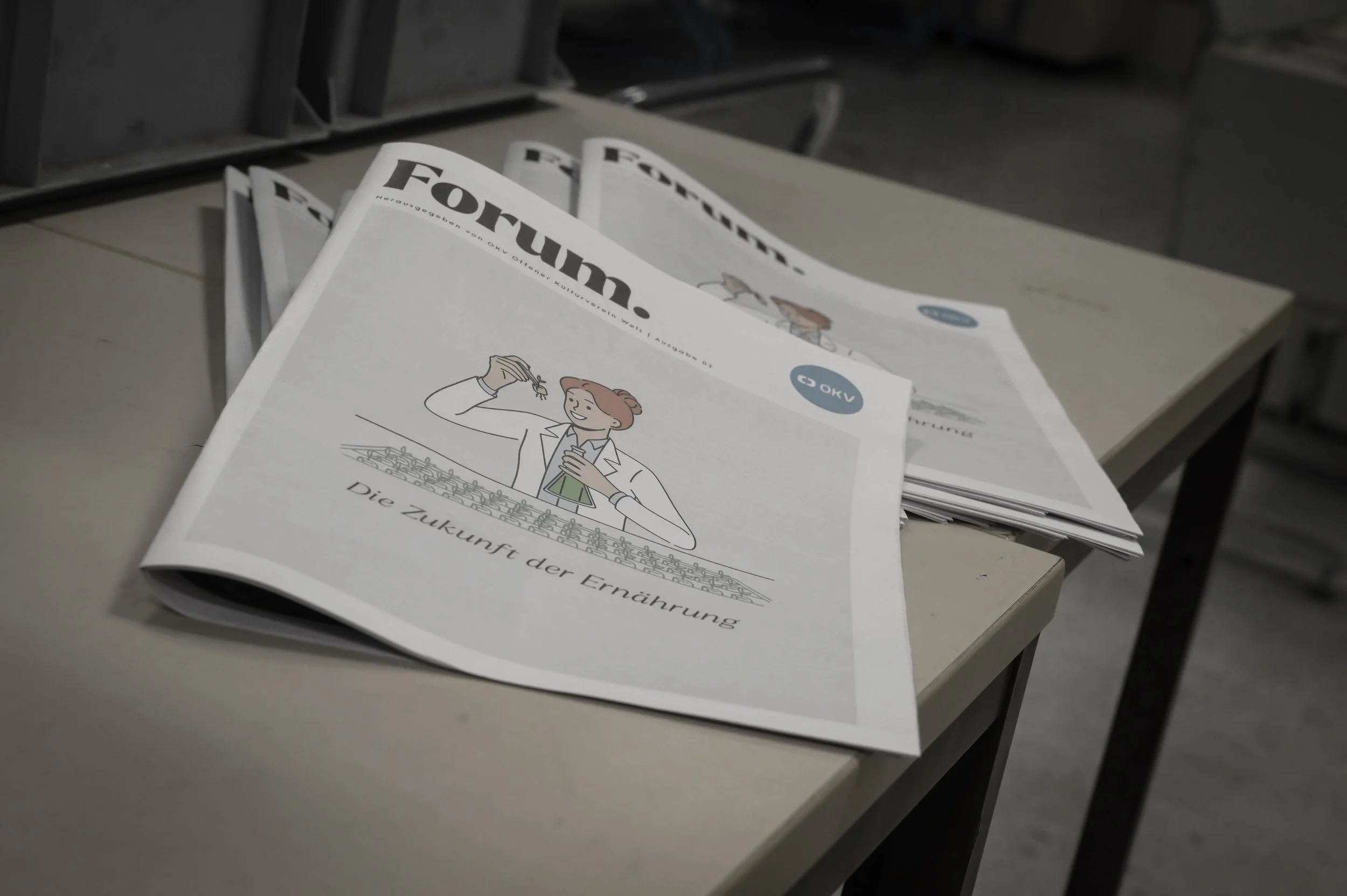 Eine Zeitung liegt auf einer weißen Oberfläche. Die Titelseite zeigt eine Illustration einer Wissenschaftlerin mit Reagenzglas, die in eine Schale mit kleinen Bildelementen schaut. Der Titel der Zeitung lautet "FOCUS" und es sind weitere Textteile un