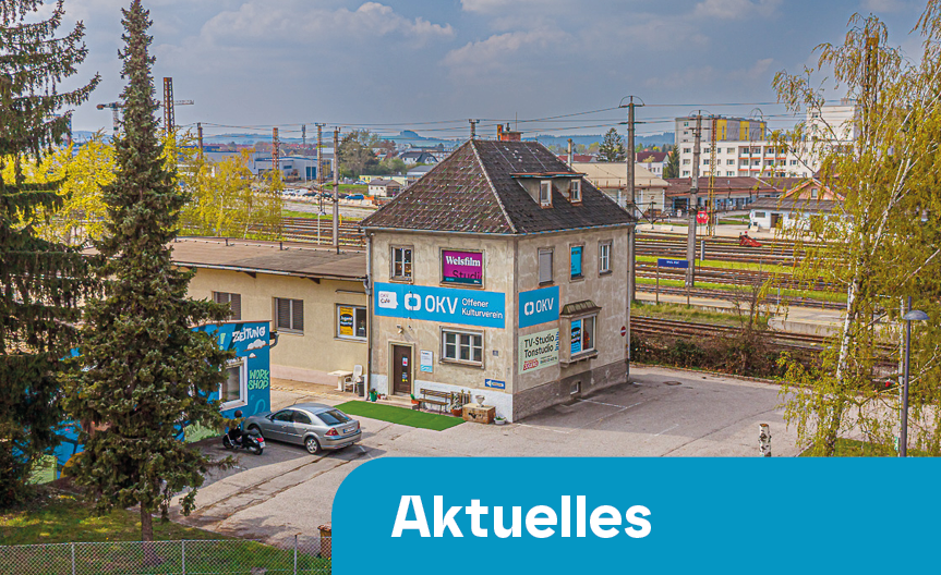 Ein zweistöckiges Gebäude mit mehreren Beschilderungen, umgeben von Bäumen und geparkten Autos, mit Bahnlinien im Hintergrund und einem blauen Banner mit dem Wort 'Aktuelles' im Vordergrund.