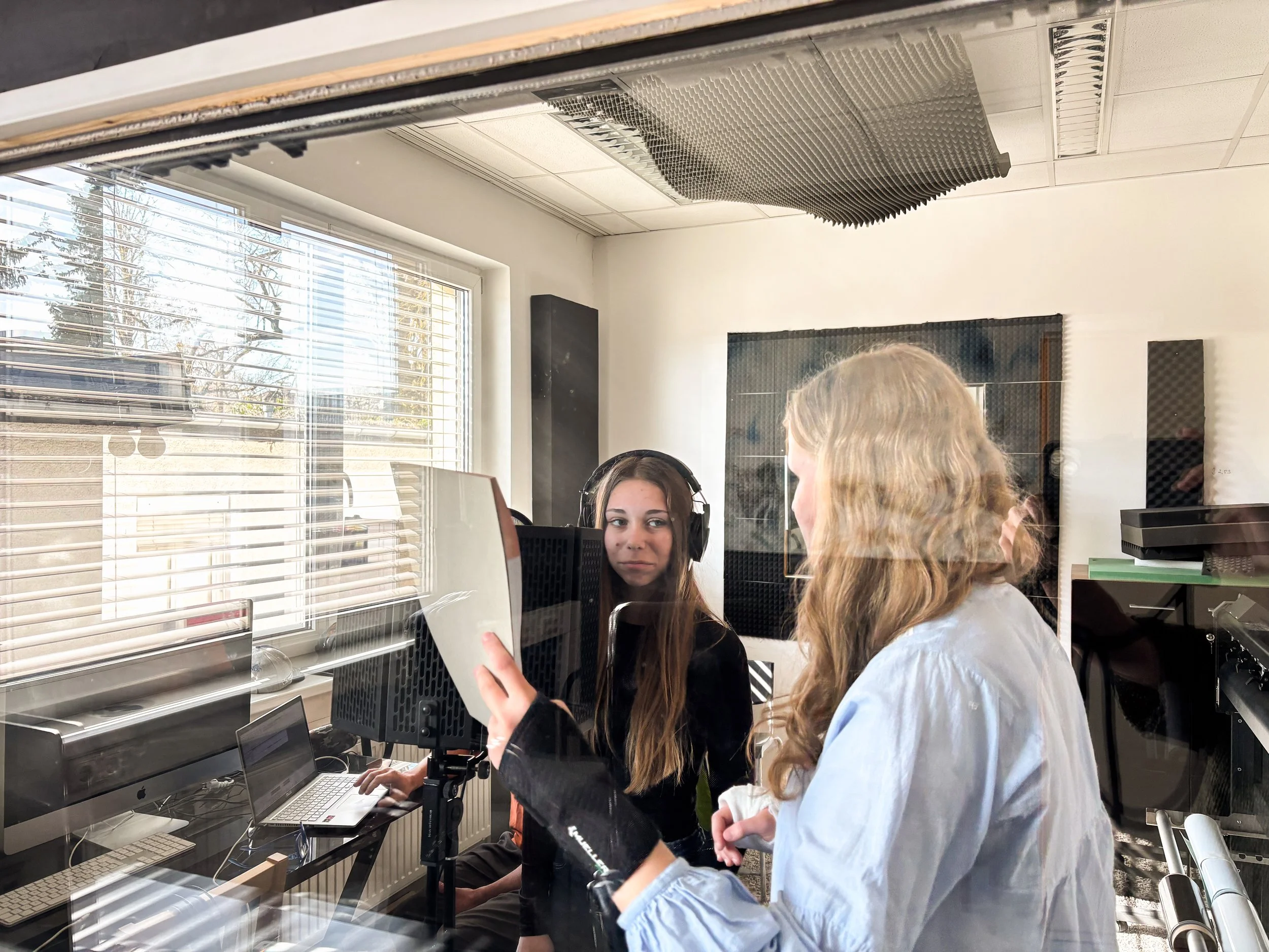 Drei Menschen, zwei junge Frauen und ein Mädchen, befinden sich in einem Raum mit Computern und Tonaufnahmeausrüstung. Sie erkunden und diskutieren die Geräte, während sie an einem Tisch neben einem Fenster stehen.