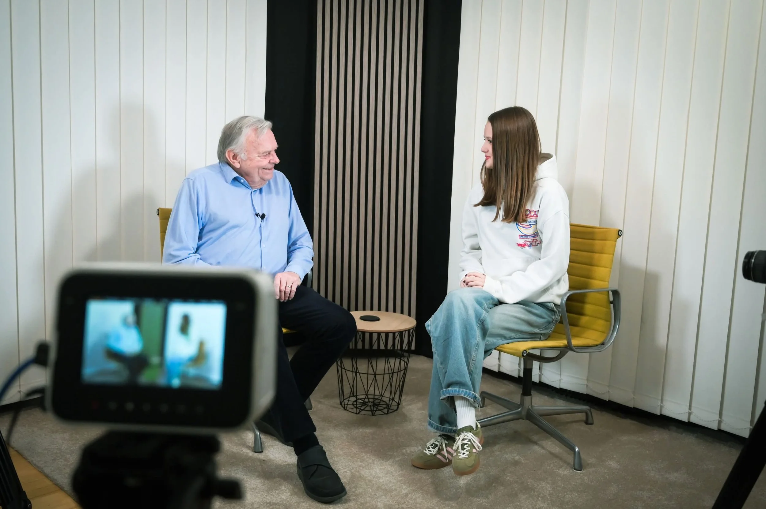 Ein älterer Mann und eine junge Frau sitzen einander gegenübersitzend in einem Raum und führen ein Gespräch. Im Vordergrund ist eine Kamera zu sehen, die das Interview aufzeichnet.