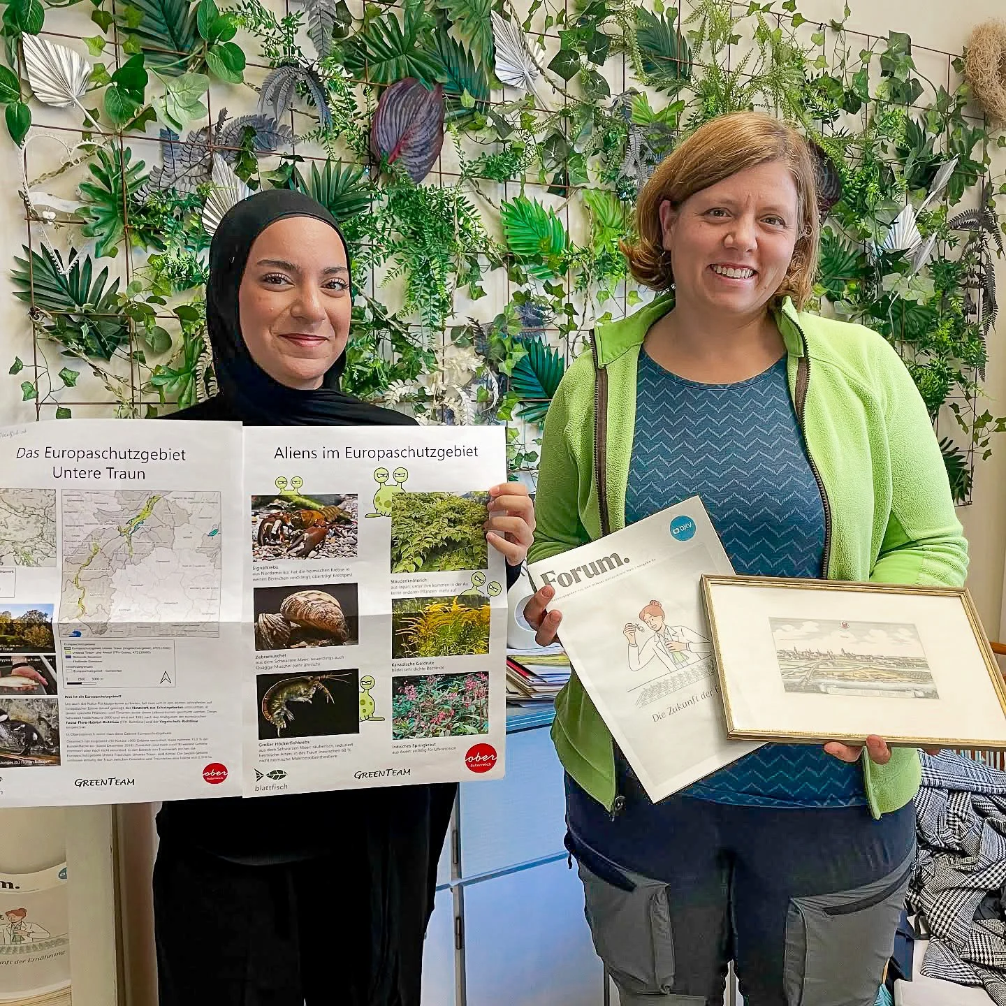 Zwei Frauen stehen vor einer grünen Pflanzenwand und halten verschiedene Informationsmaterialien in den Händen. Die Frau auf der linken trägt einen schwarzen Hijab, die Frau auf der rechten trägt eine grüne Sweatjacke.