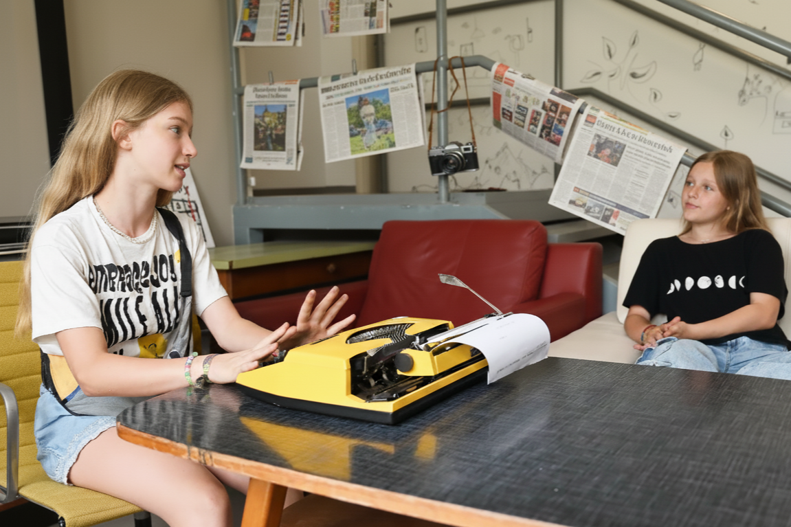 Zwei Mädchen unterhalten sich in einem Raum, einer sitzt am Tisch mit einer Schreibmaschine, der andere sitzt auf einem Sofa. Im Hintergrund hängen Zeitungsausschnitte und es ist eine Kamera sichtbar.