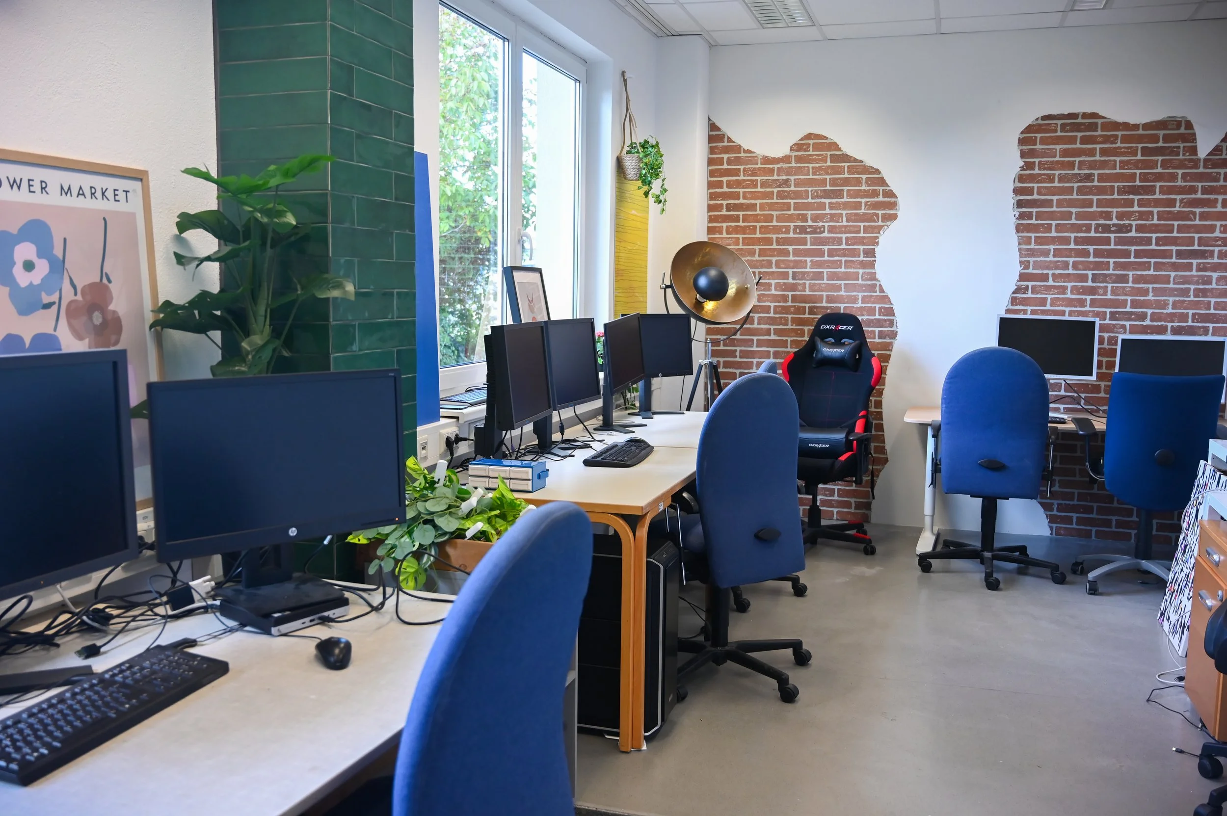 Offene Büro-Umgebung mit mehreren Computern, blauen Stühlen, grünen Pflanzen, einem roten Gaming-Stuhl und einer Wand mit teilweise abgeblättertem Ziegelmuster.