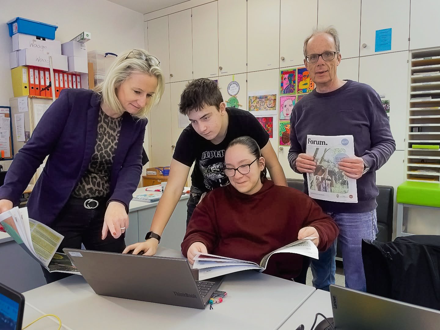 Vier Personen arbeiten um einen Laptop in einem Klassenzimmer mit bunten Kunstwerken an der Wand.