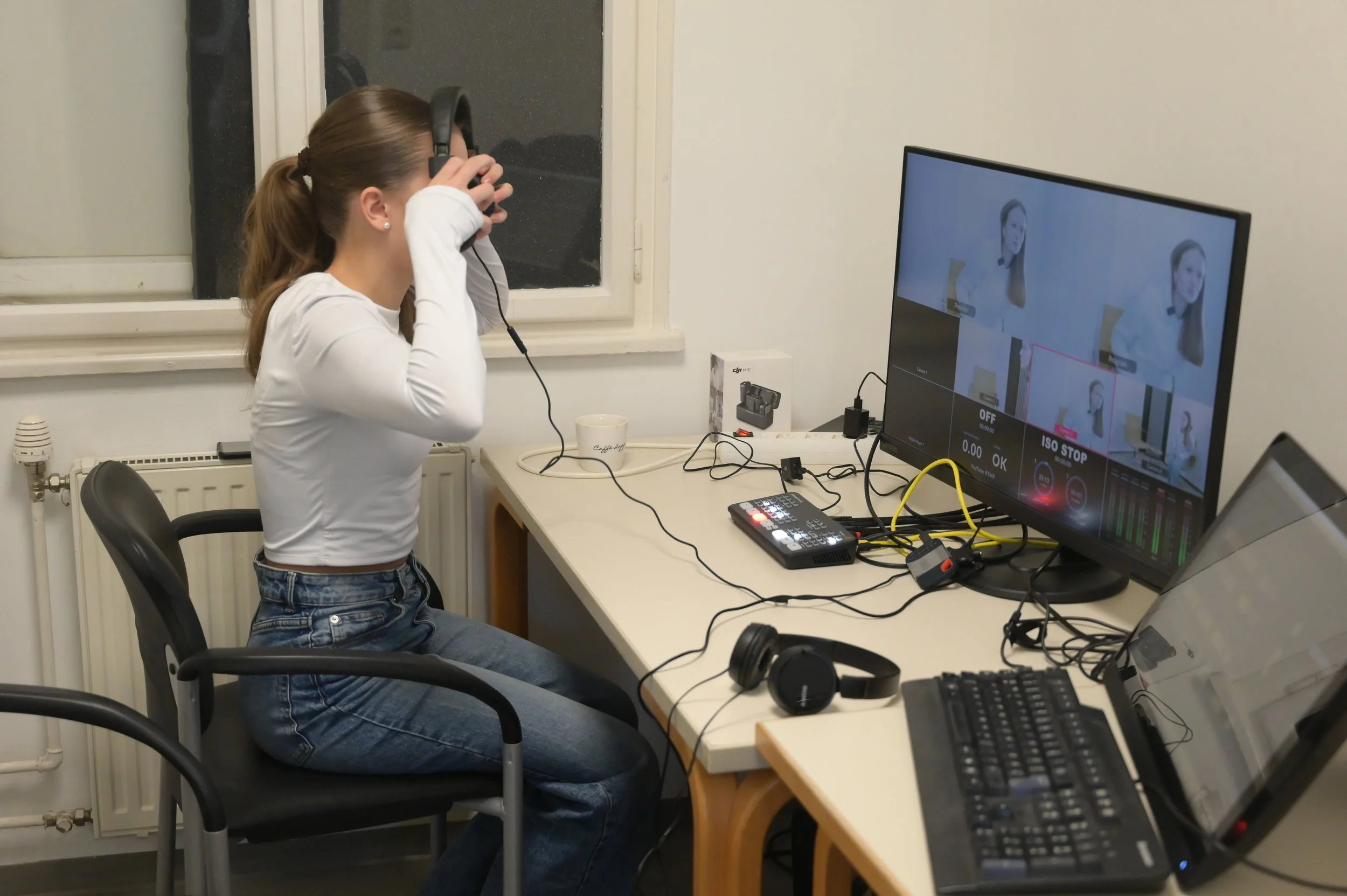 Eine junge Frau sitzt an einem Schreibtisch, trägt Kopfhörer und schaut auf einen Computerbildschirm, auf dem mehrere Bilder einer Frau zu sehen sind.