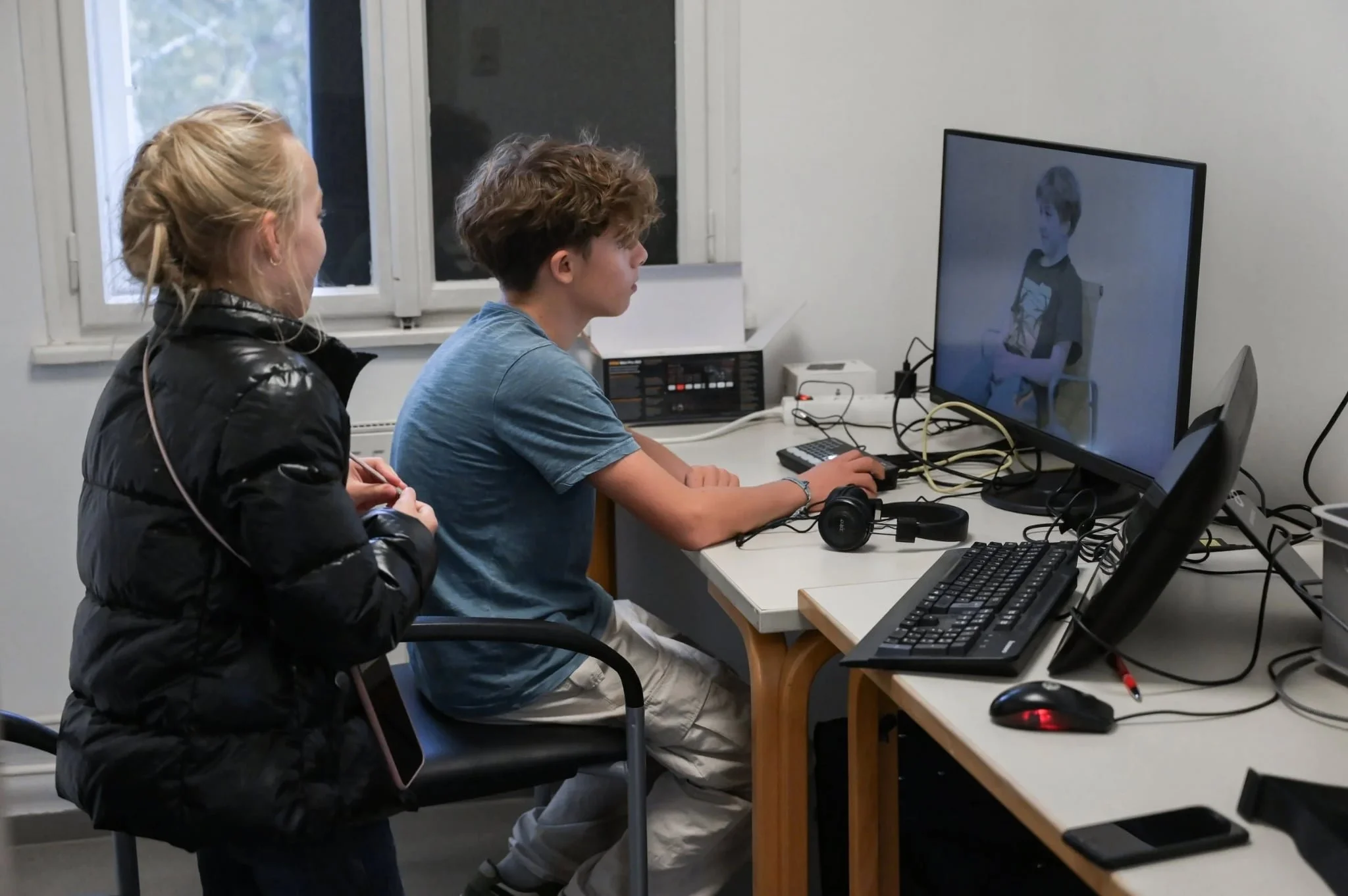 Ein Teenager sitzt an einem Schreibtisch und spielt ein Computerspiel auf einem großen Monitor, während eine Frau neben ihm steht und zuschaut.