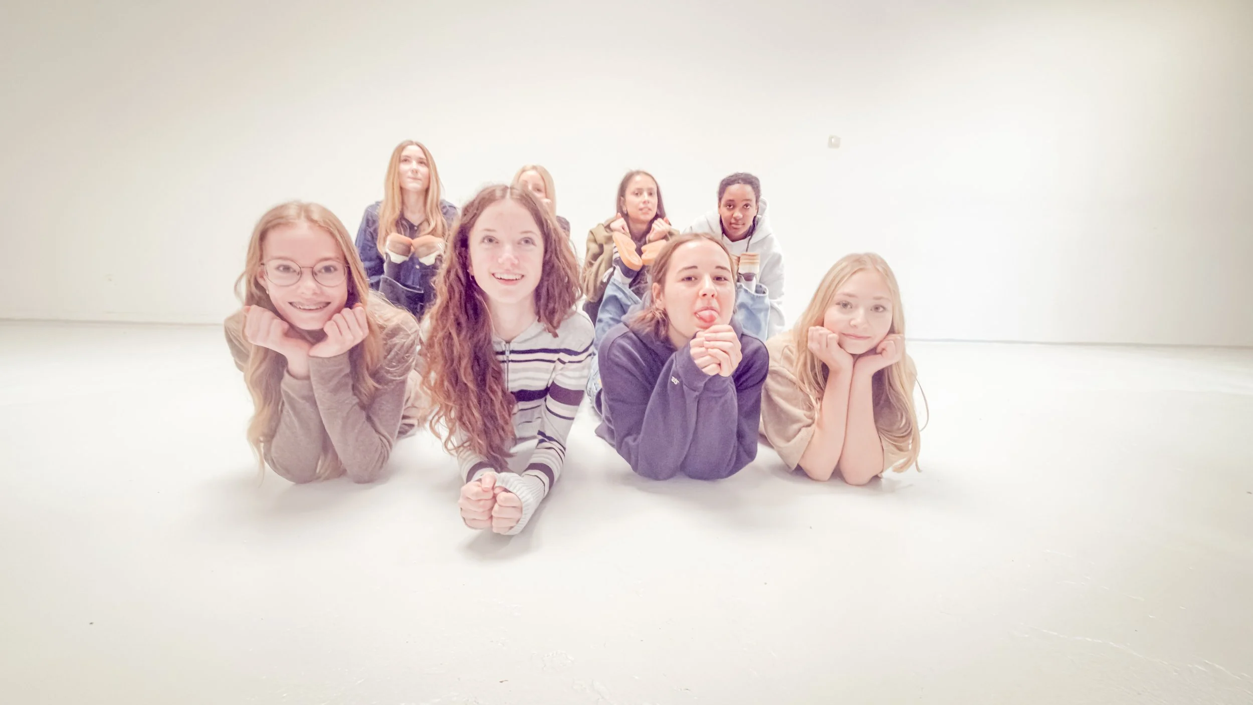 Eine Gruppe von sieben jungen Frauen liegt auf dem Boden in einem weißen Raum, posiert für ein Foto und wirkt entspannt und fröhlich.