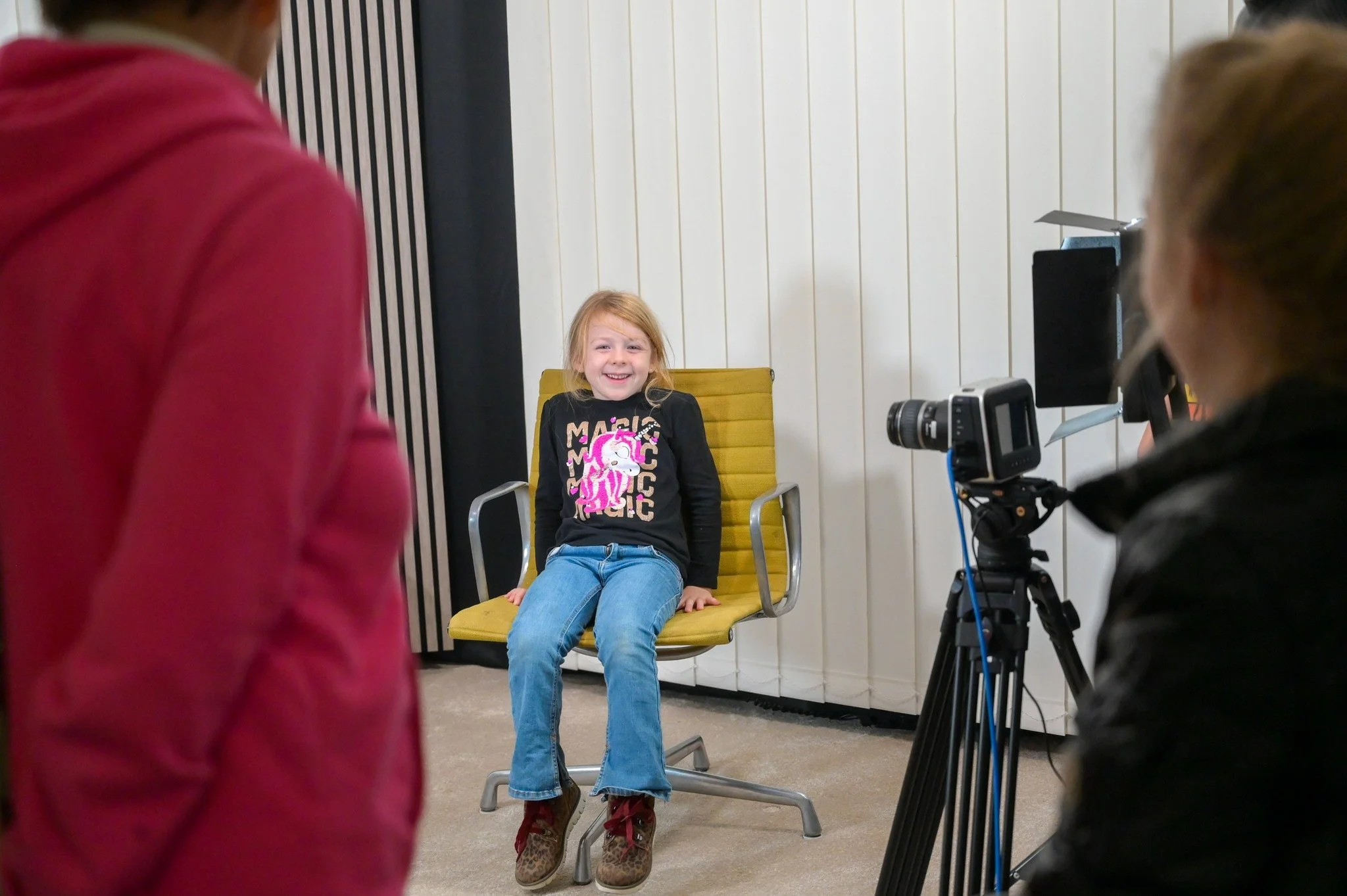 Ein Mädchen sitzt lachend auf einem Stuhl während eines Fotoshootings, im Hintergrund eine weiße Wand mit vertikalen schwarzen Linien, umgeben von Personen, die fotografieren und filmen.