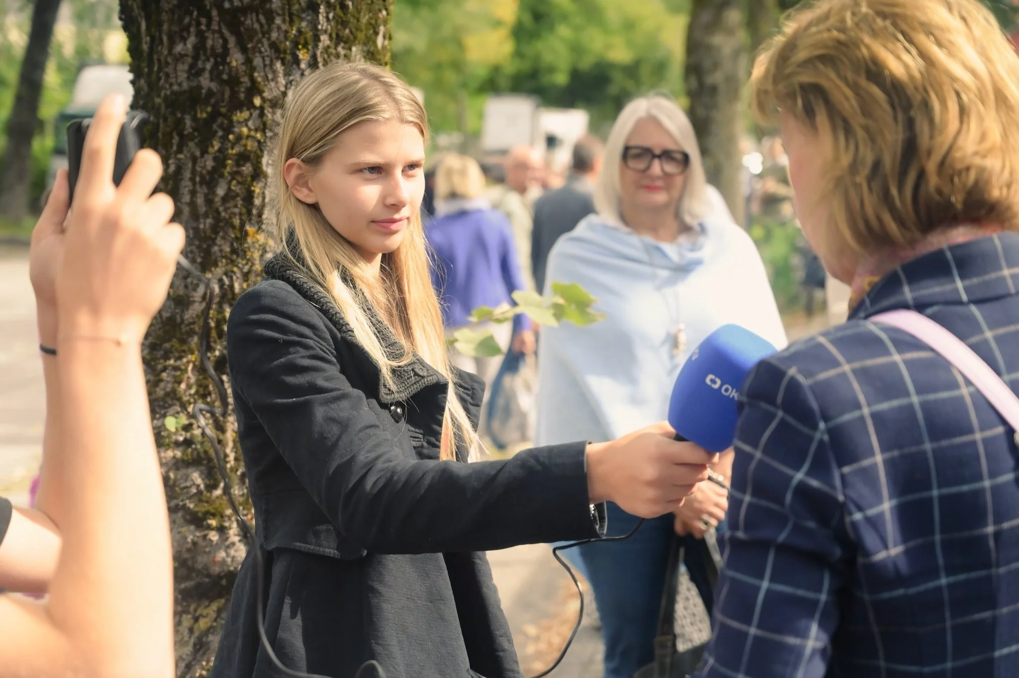 Junge Frau wird von einer Reporterin interviewt, im Hintergrund stehen mehrere Personen bei einem Außenereignis