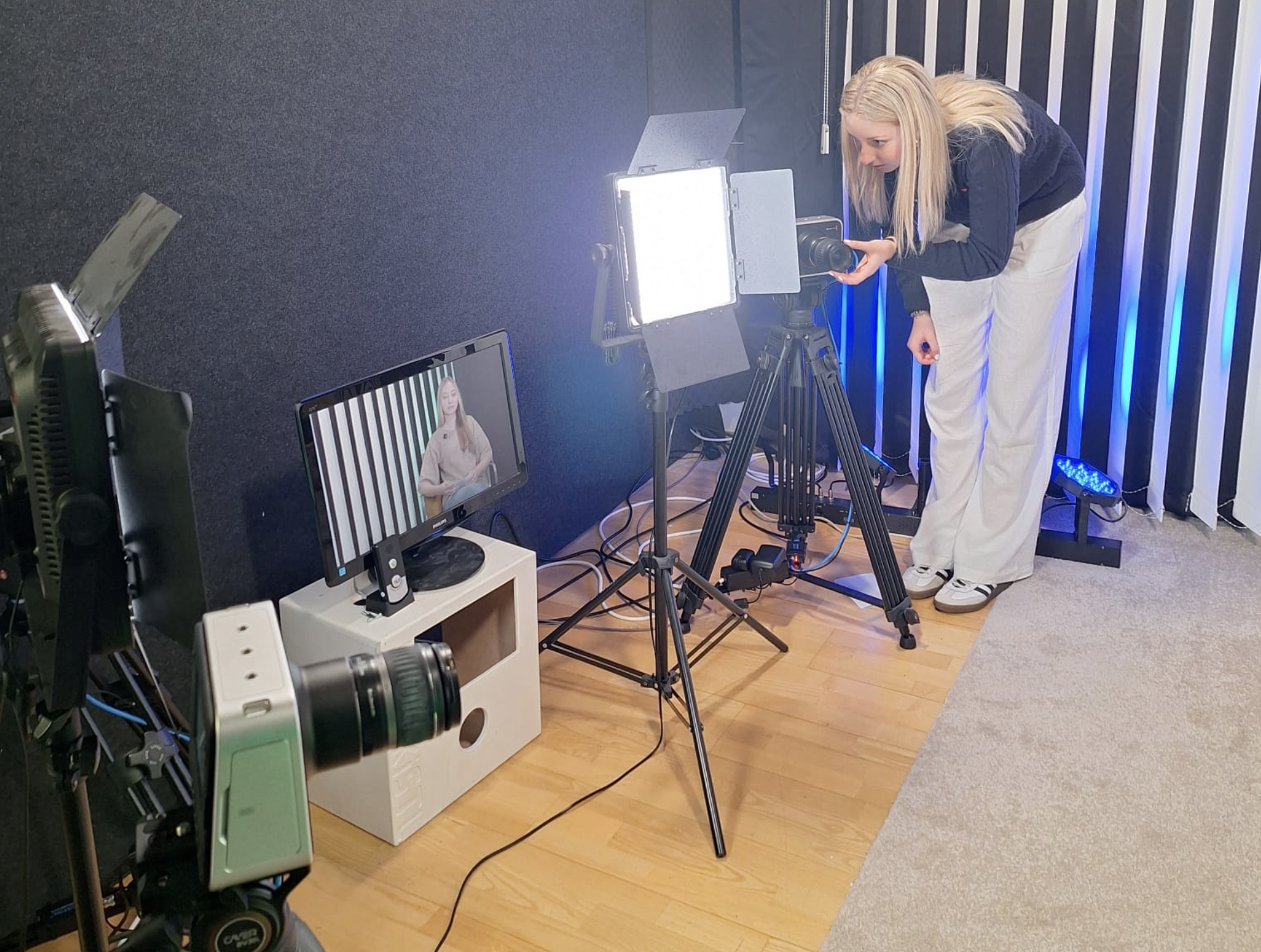 Eine Frau wird in einem Studio für eine Videoproduktion gefilmt, mit professioneller Kamera und Lichtquelle, ein Monitor zeigt die Aufnahme eines Videos. Der Raum hat eine dunkle Wand mit vertikalen weißen Linien.