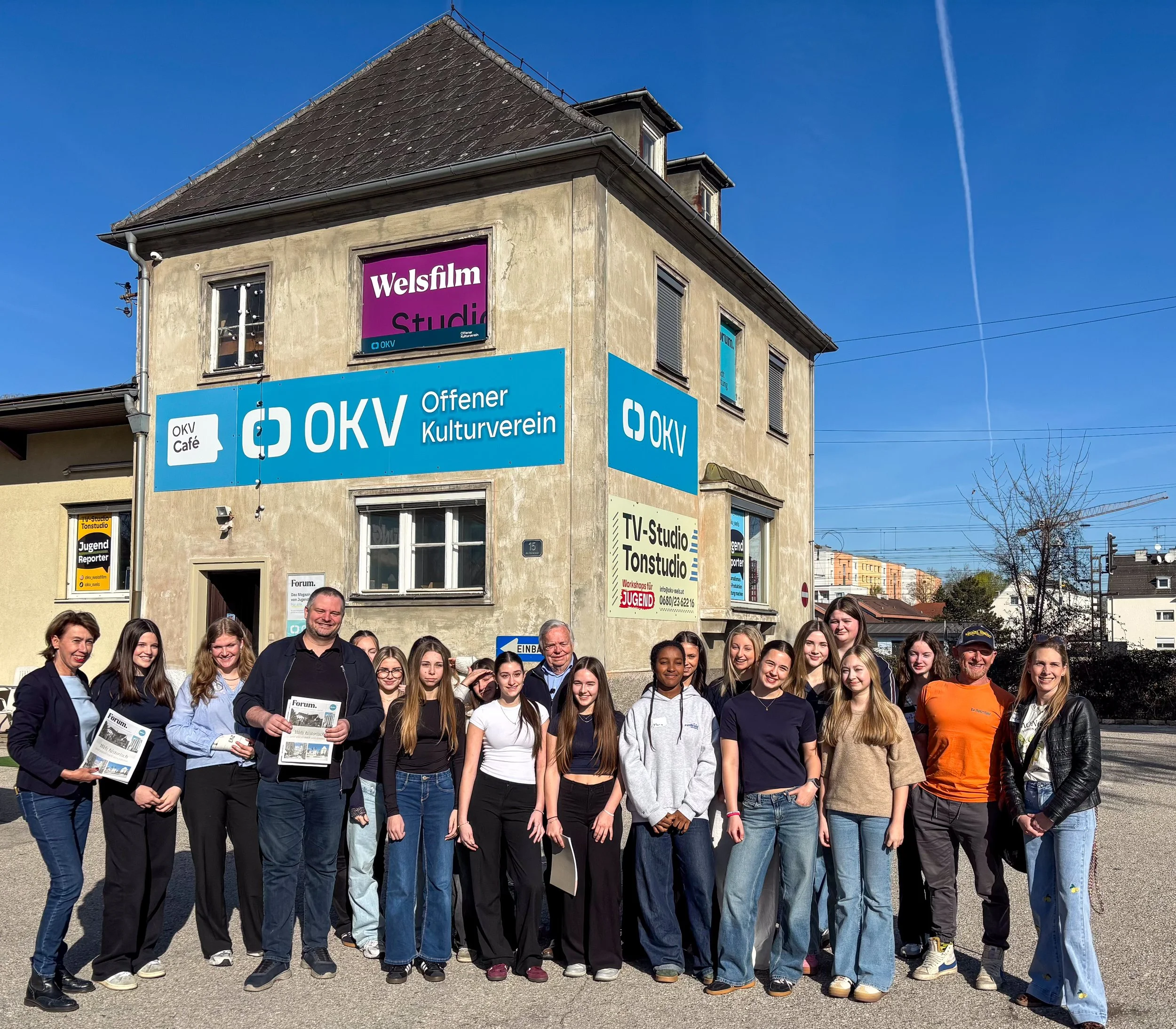 Gruppe von Menschen vor einem Gebäude mit blauen OKV-Bannern, die sich für ein gemeinsames Foto versammelt haben, bei sonnigem Wetter in einer Stadt mit mehreren Gebäuden im Hintergrund.