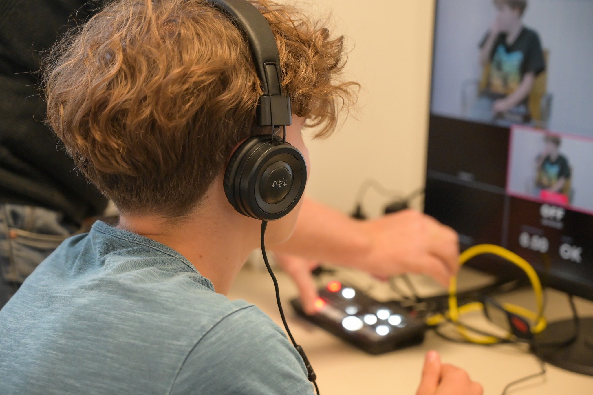Junge Person mit lockigem Haar trägt Kopfhörer und arbeitet an einem DJ- oder Musikgerät, im Hintergrund ein Bildschirm mit einem Bild eines Mädchens.