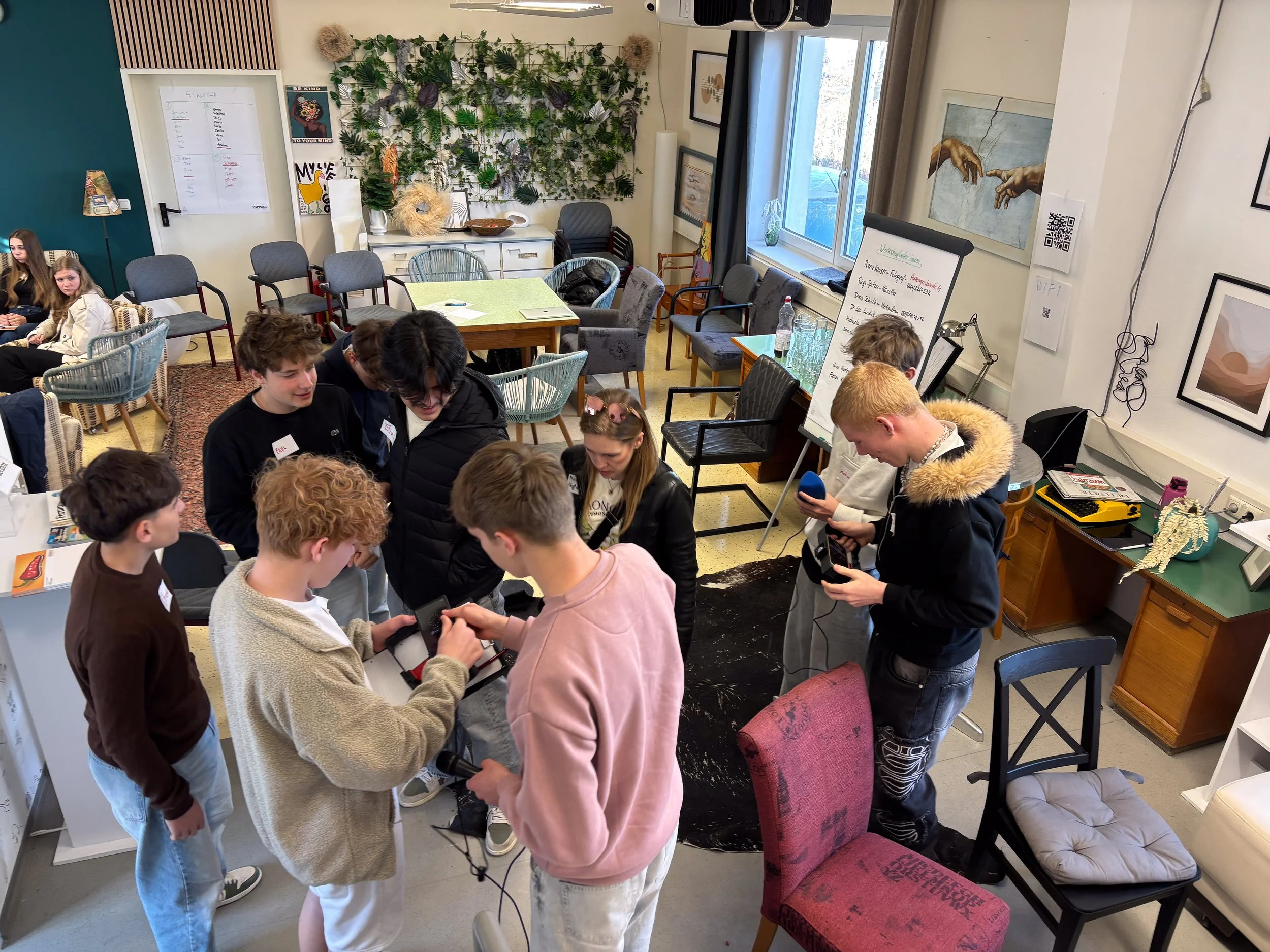 Gruppen junger Menschen, die in einem Raum zusammenstehen und mit Mobiltelefonen interagieren, während andere im Hintergrund sitzen und sich unterhalten.