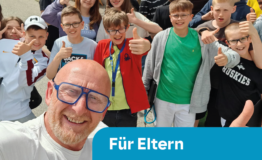 Lächelnder Mann mit blauen Brillen vor einer Gruppe fröhlicher Kinder, die Daumen hoch zeigen, im Freien bei gutem Wetter.