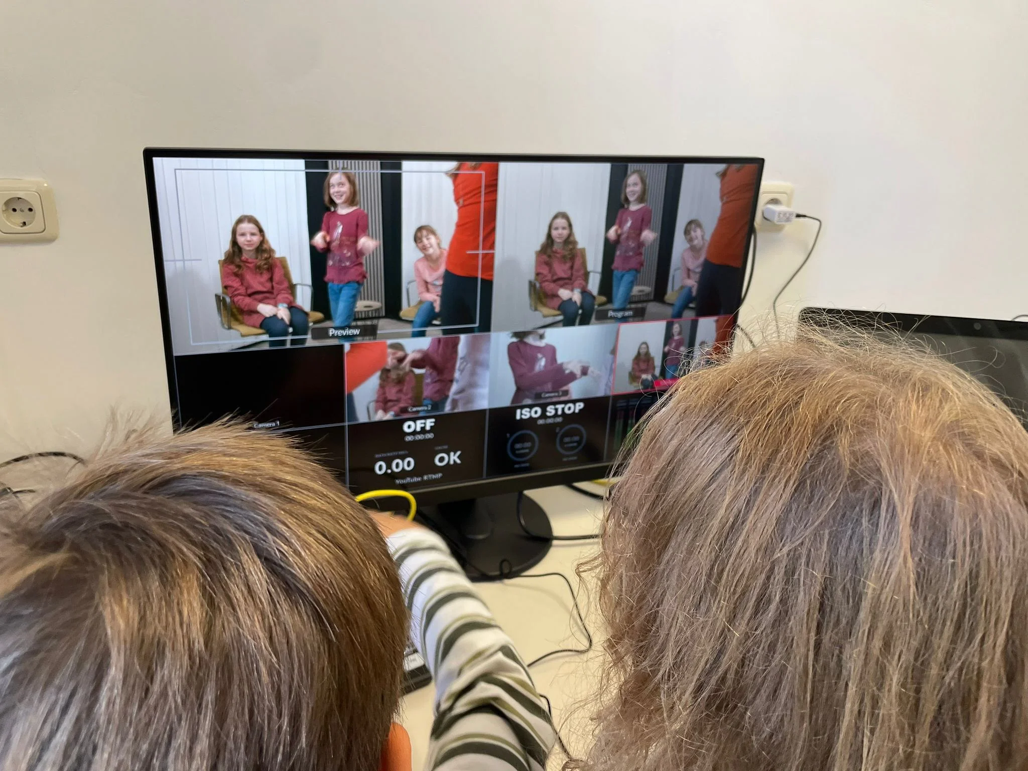 Auf einem Bildschirm sieht man eine Gruppe von Kinder, die auf Stühlen sitzen, während jemand in Orange vor ihnen steht. Der Bildschirm zeigt mehrere Kameraperspektiven des Kindes und der Person, die spricht.