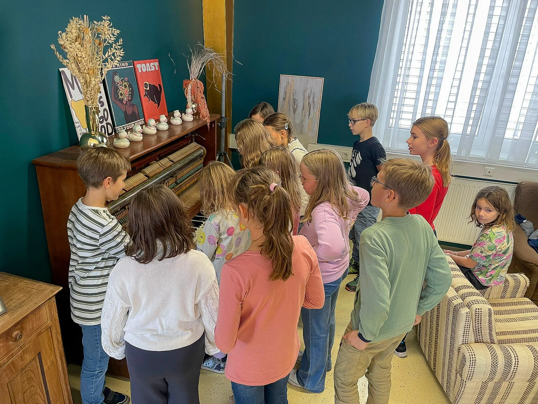 Gruppen von Kindern und Jugendlichen stehen in einem Raum um einen alten Flügel und hören zu, während eine Person den Flügel spielt.