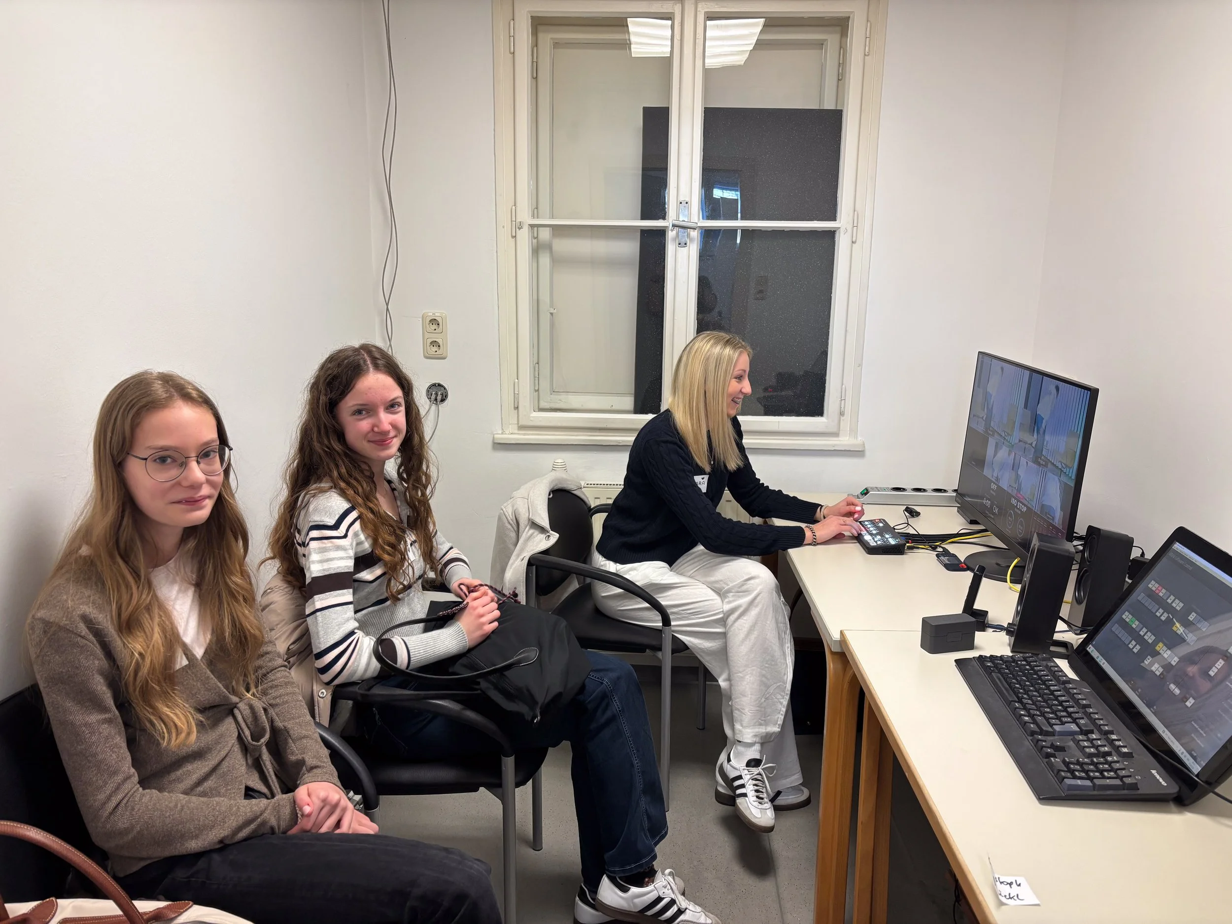 Vier Frauen sitzen in einem Büro um einen Tisch, auf dem Computer und Monitore stehen. Eine Frau arbeitet am Computer, die anderen drei schauen in die Kamera, alle lächeln.
