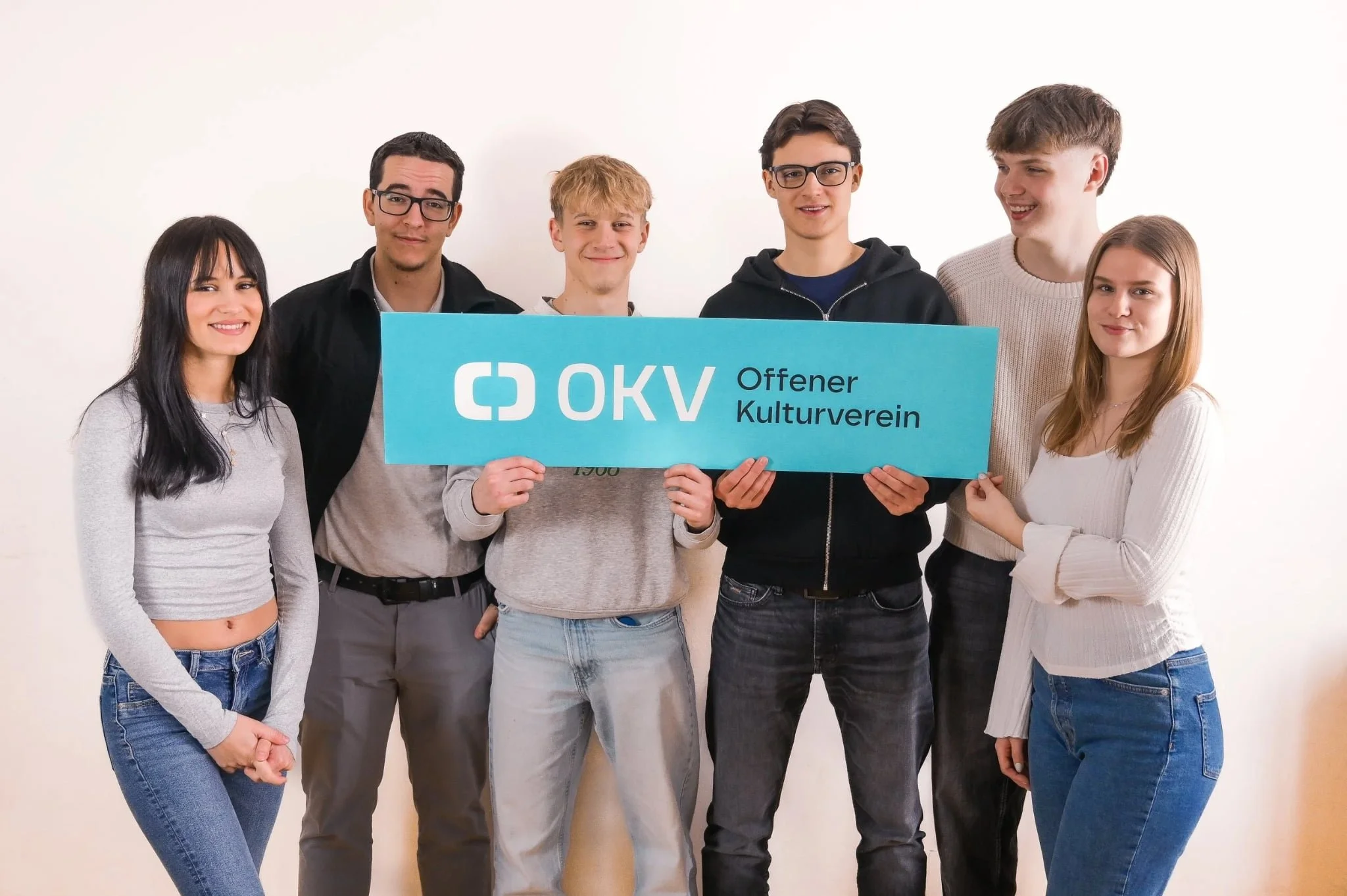 Sechs junge Menschen stehen zusammen und halten ein Helen a blaues Schild mit dem Text "OKV Offener Kulturverein" vor einem weißen Hintergrund, lächeln und schauen in die Kamera.
