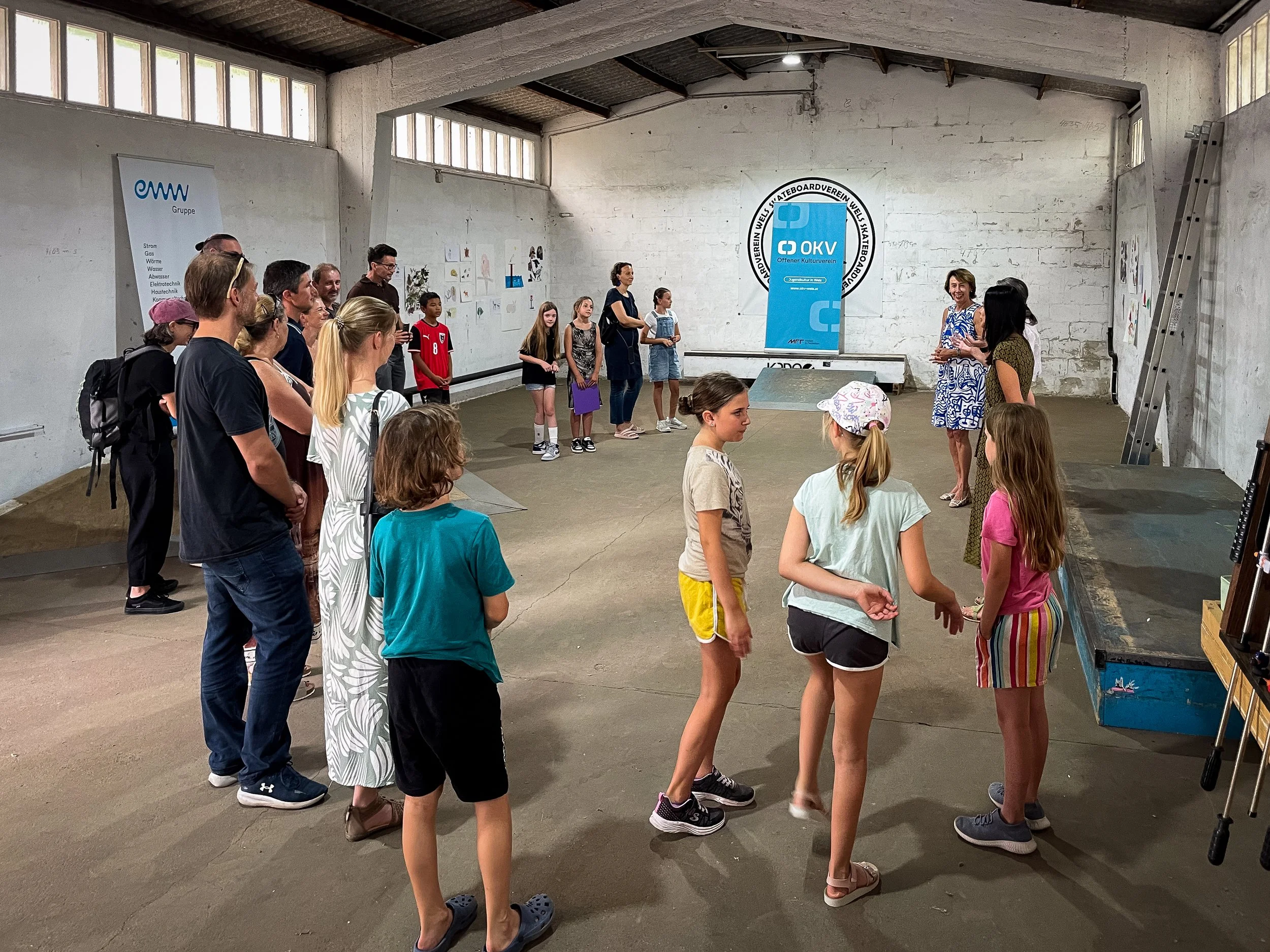 Gruppen von Kindern und Erwachsenen in einem Raum, die einer Lehrerin zuhören, vor einer Pinnwand und einem Banner mit dem Logo des Skatervereins Wels. Es gibt Wandzeichnungen und eine Leiter an der Wand.