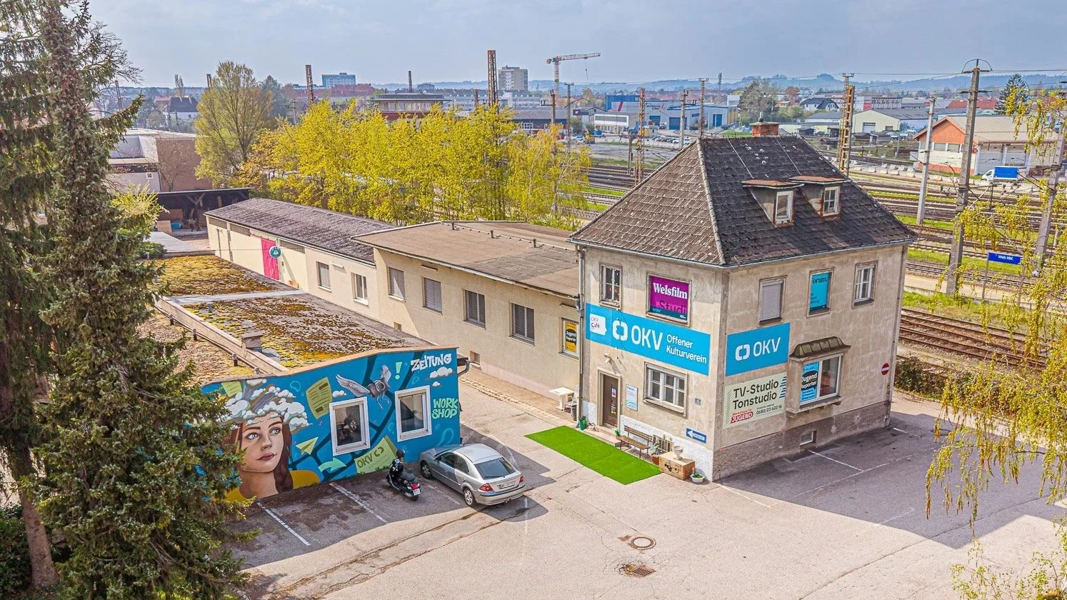 Ein Mehrfamilienhaus mit Werbetafeln und Graffiti vor einem Parkplatz, umgeben von Bäumen und einer Eisenbahnstrecke im Hintergrund.