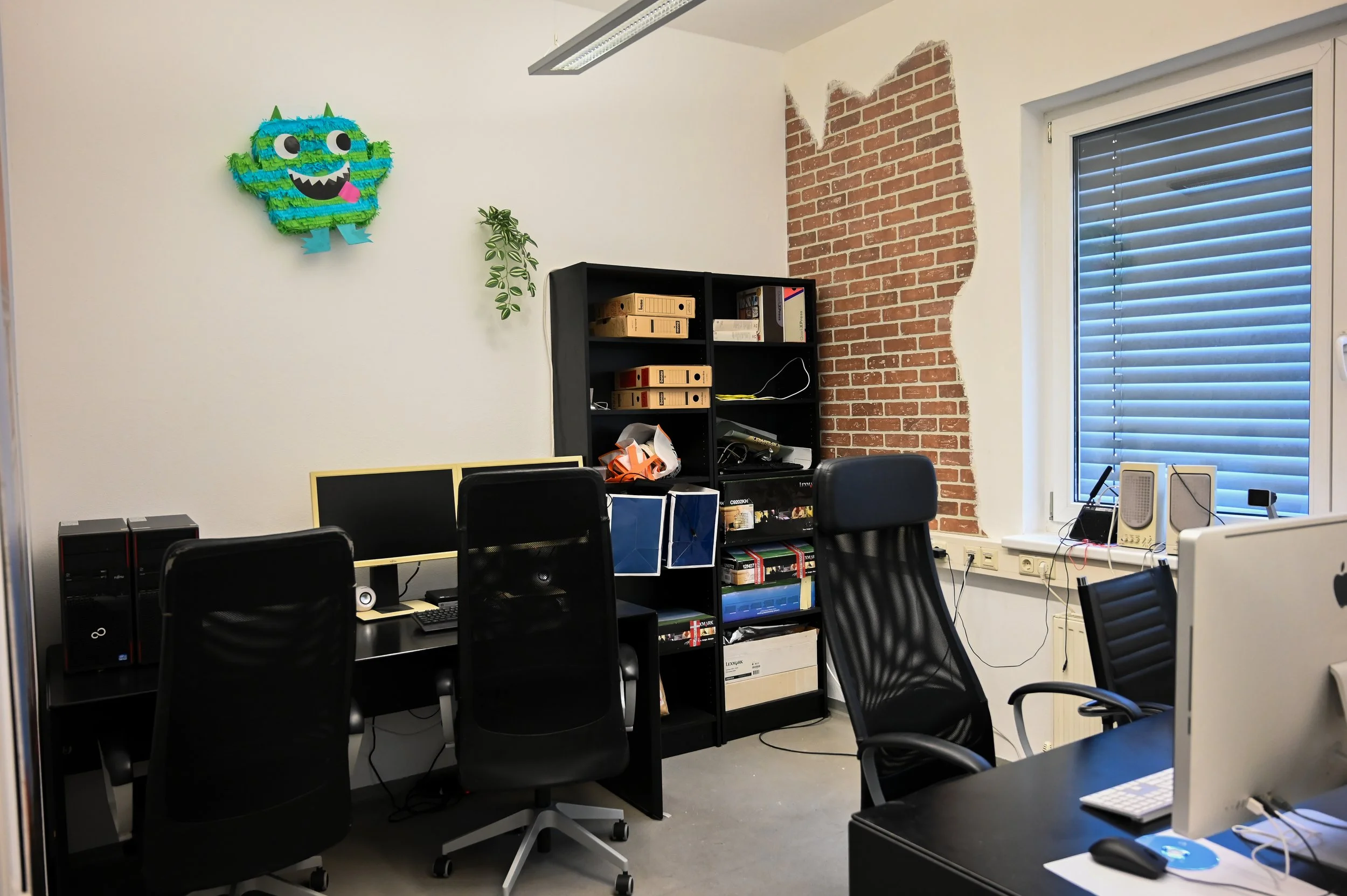 Büro mit Schreibtischen, schwarzen Stühlen, einem Regal mit Ordnern, einer Dekoration einer grünen, blauen und lila Piñata in Form eines Monsters an der Wand, und einer großen Fensterbrüstung mit Rollläden.