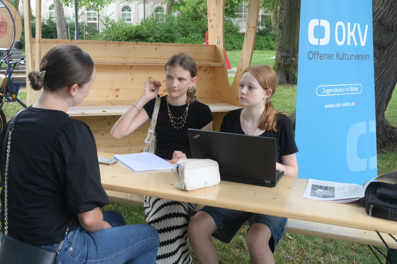 Drei junge Mädchen sitzen an einem Holztisch im Freien, gegenüber einer Frau, die sie interviewt. Im Hintergrund ist ein blauer Stand mit dem Logo des Offenen Kulturvereins (OKV) und einem Schild mit der Aufschrift ‚Jugendkultur in Wels‘ sichtbar.