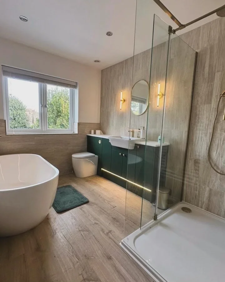 Modern bathroom with a free-standing bathtub, a toilet, a green vanity with a sink, a round mirror, a glass shower enclosure, a window with a view of trees, and wood-style flooring.